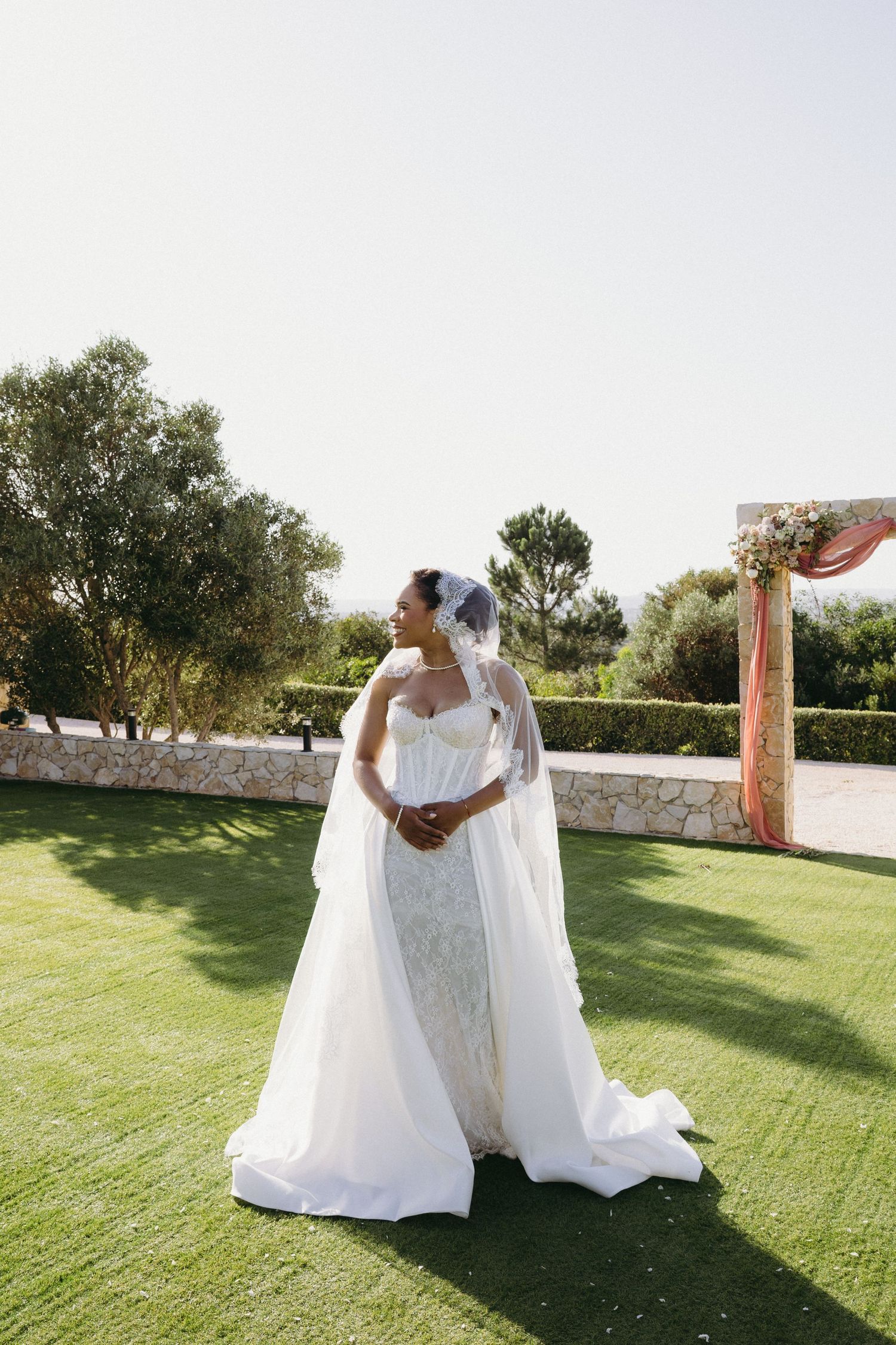 Elegant bride in white strapless gown and long veil stands on green lawn at outdoor wedding venue on sunny day.