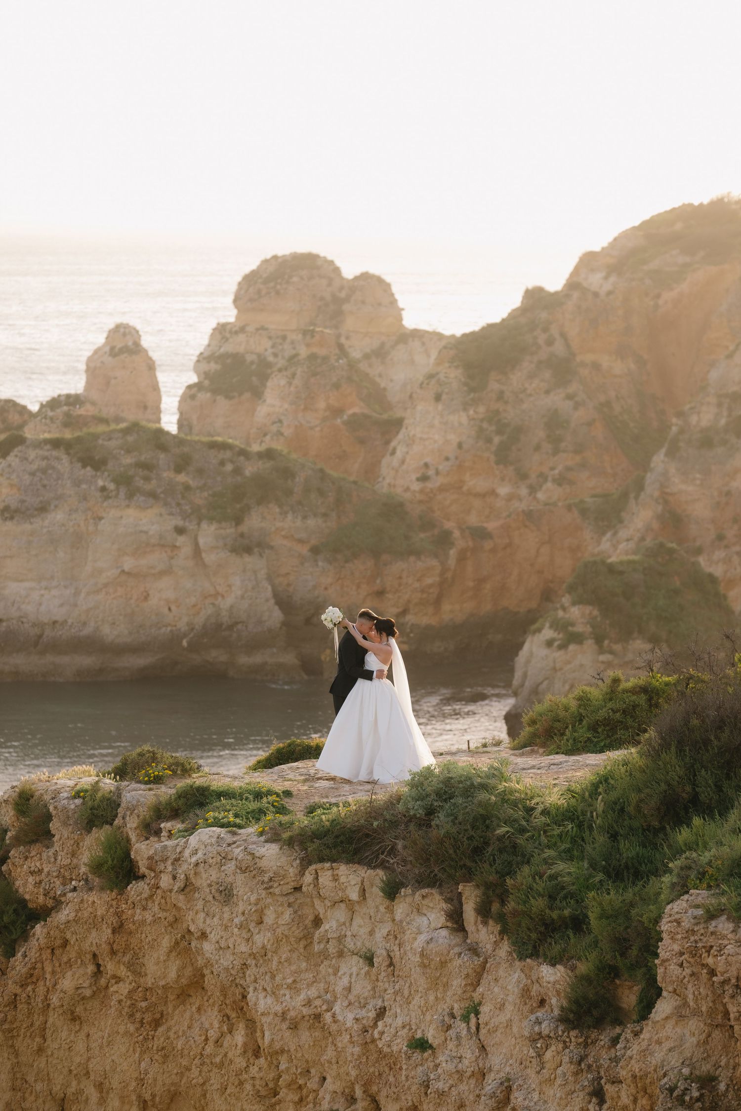 Bride and groom embracing on rocky coastal cliffs at sunset with ocean views behind them.