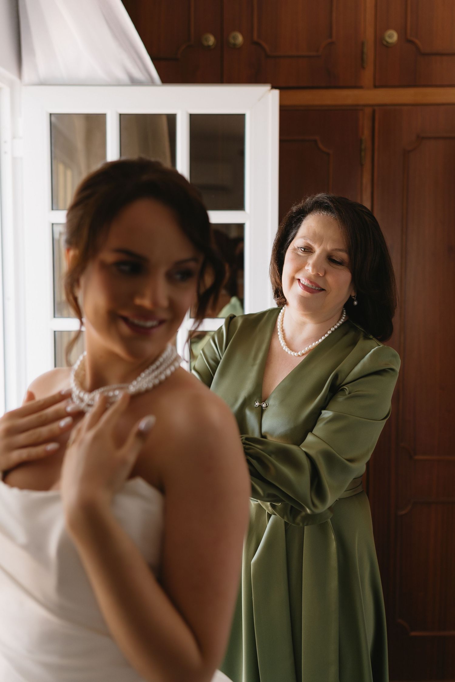 Mother in green dress smiling as she helps bride put on pearl necklace before wedding ceremony.