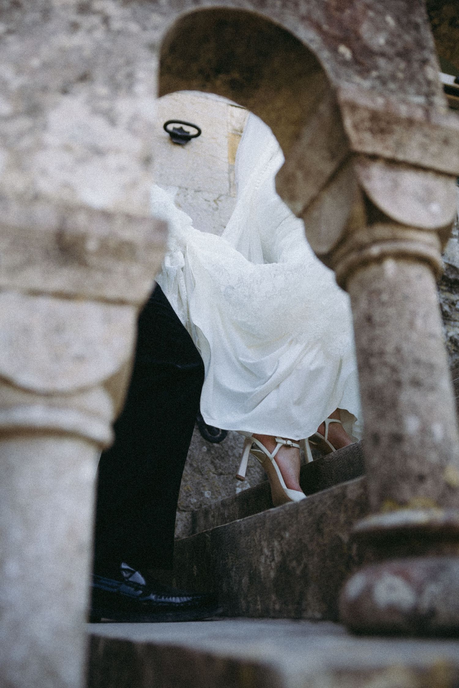 Bride in white wedding dress and veil ascending stone steps through ancient arched doorway.