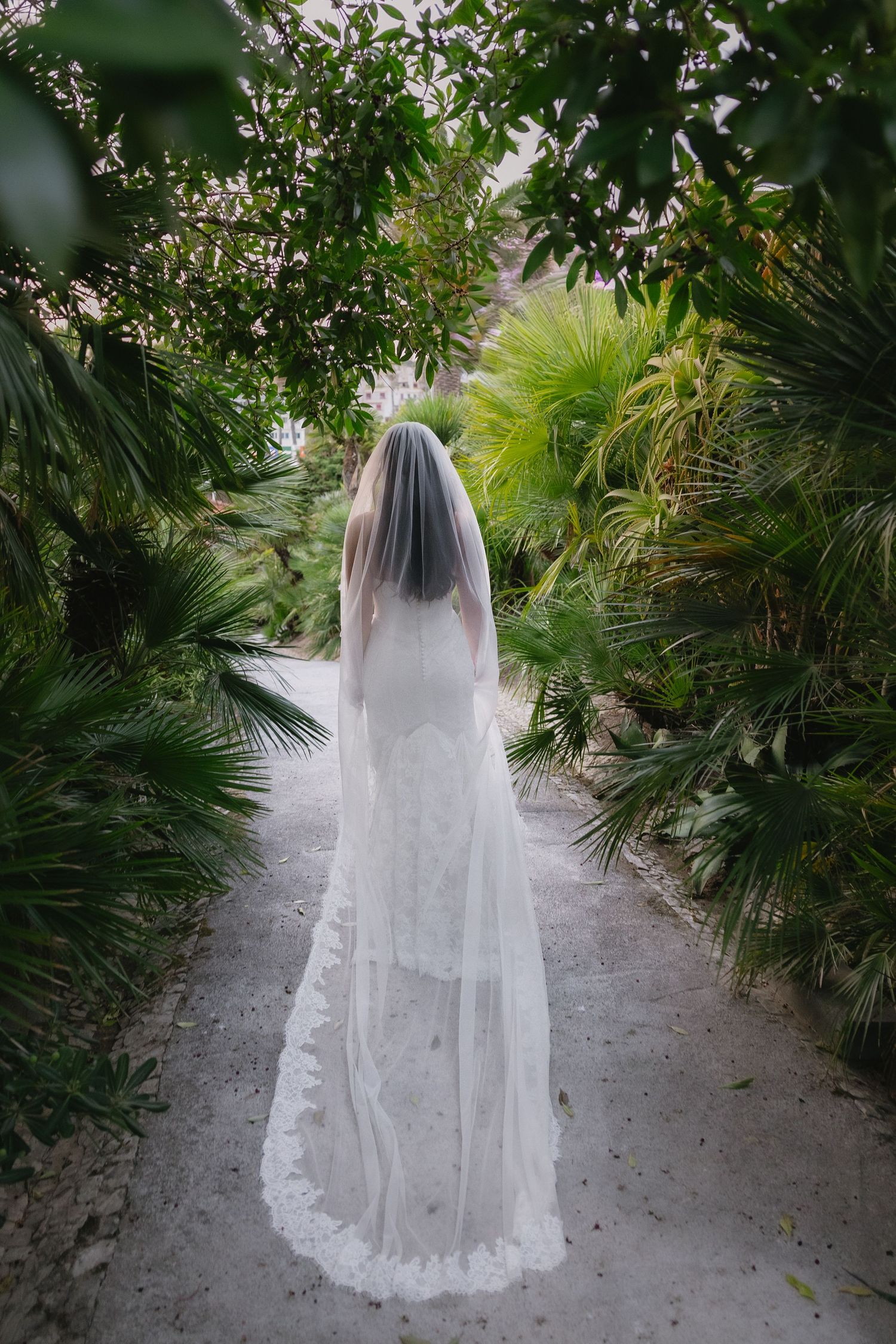 Bride in white lace wedding gown and cathedral veil walks through lush tropical garden path.