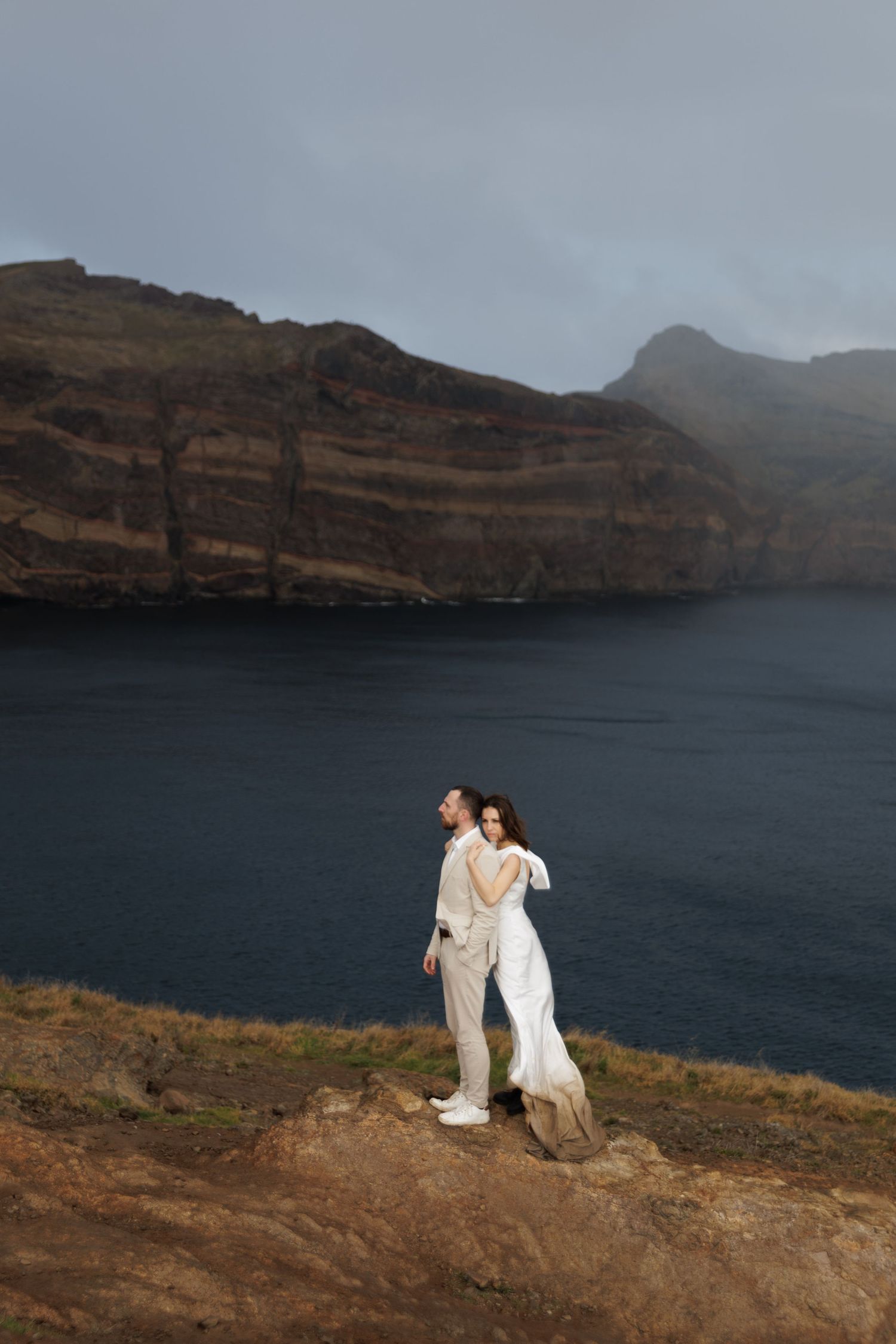 Couple in white wedding attire standing on rocky cliff overlooking dramatic ocean bay.