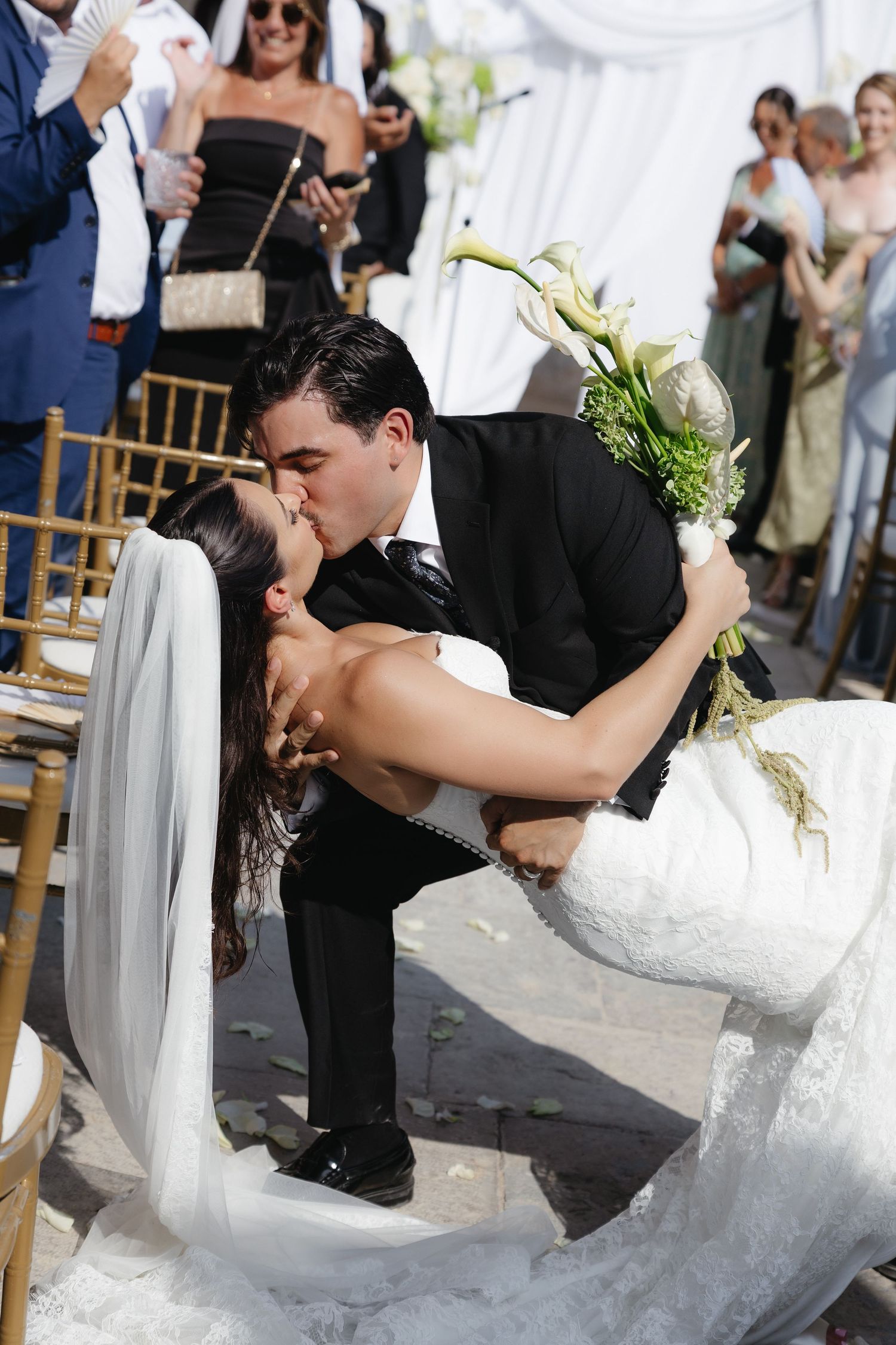 Bride and groom share a romantic dip kiss at outdoor wedding ceremony, bride in lace gown holding white calla lily bouquet.