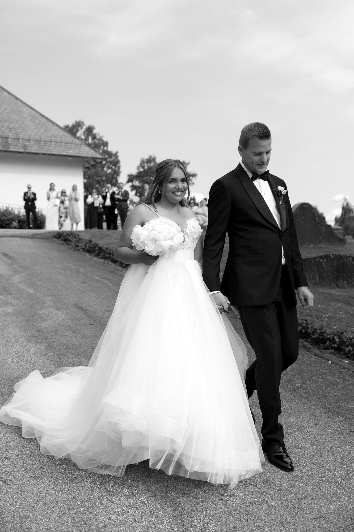 Black and white photo of bride in ball gown and groom in tuxedo walking together on path at outdoor wedding venue.