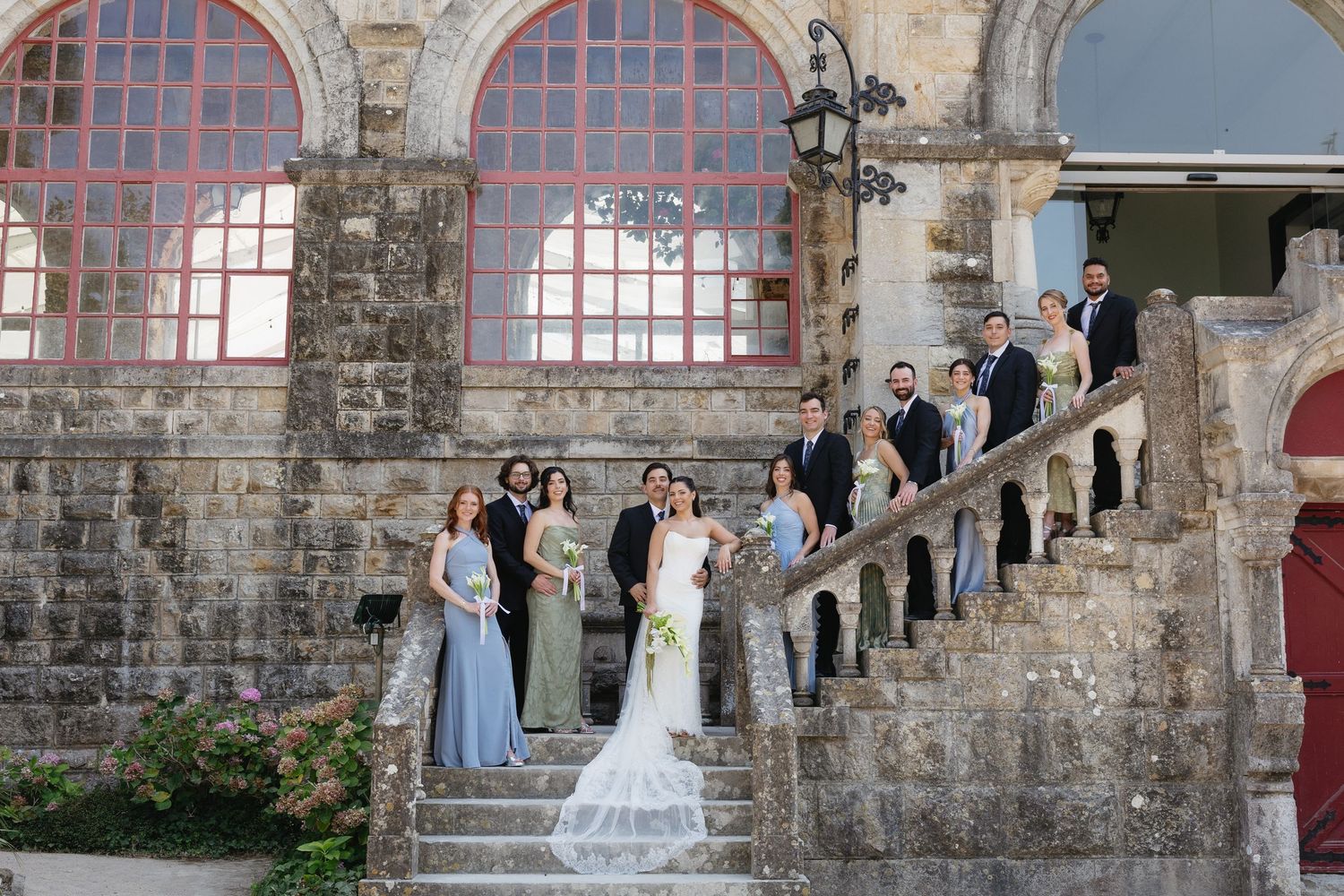Wedding party poses on stone staircase of historic venue, bride in white gown with bridesmaids in blue dresses.
