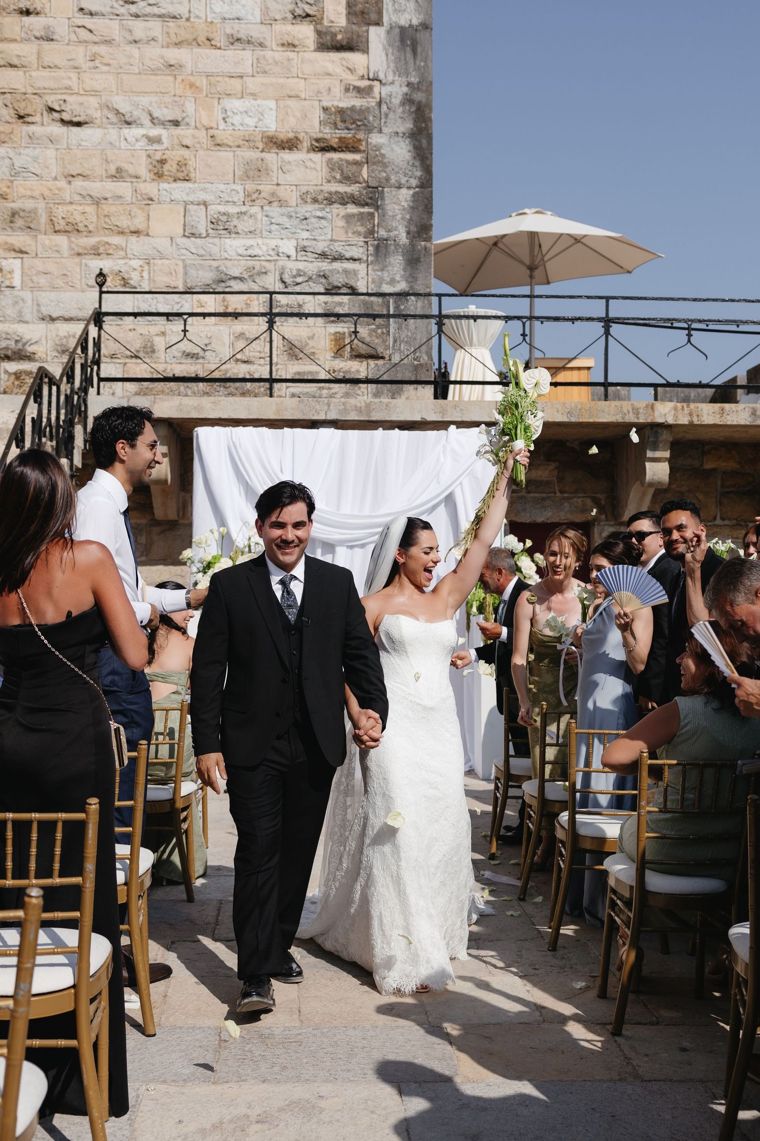 Bride and groom walk down outdoor aisle after wedding ceremony, bride raising bouquet in celebration.