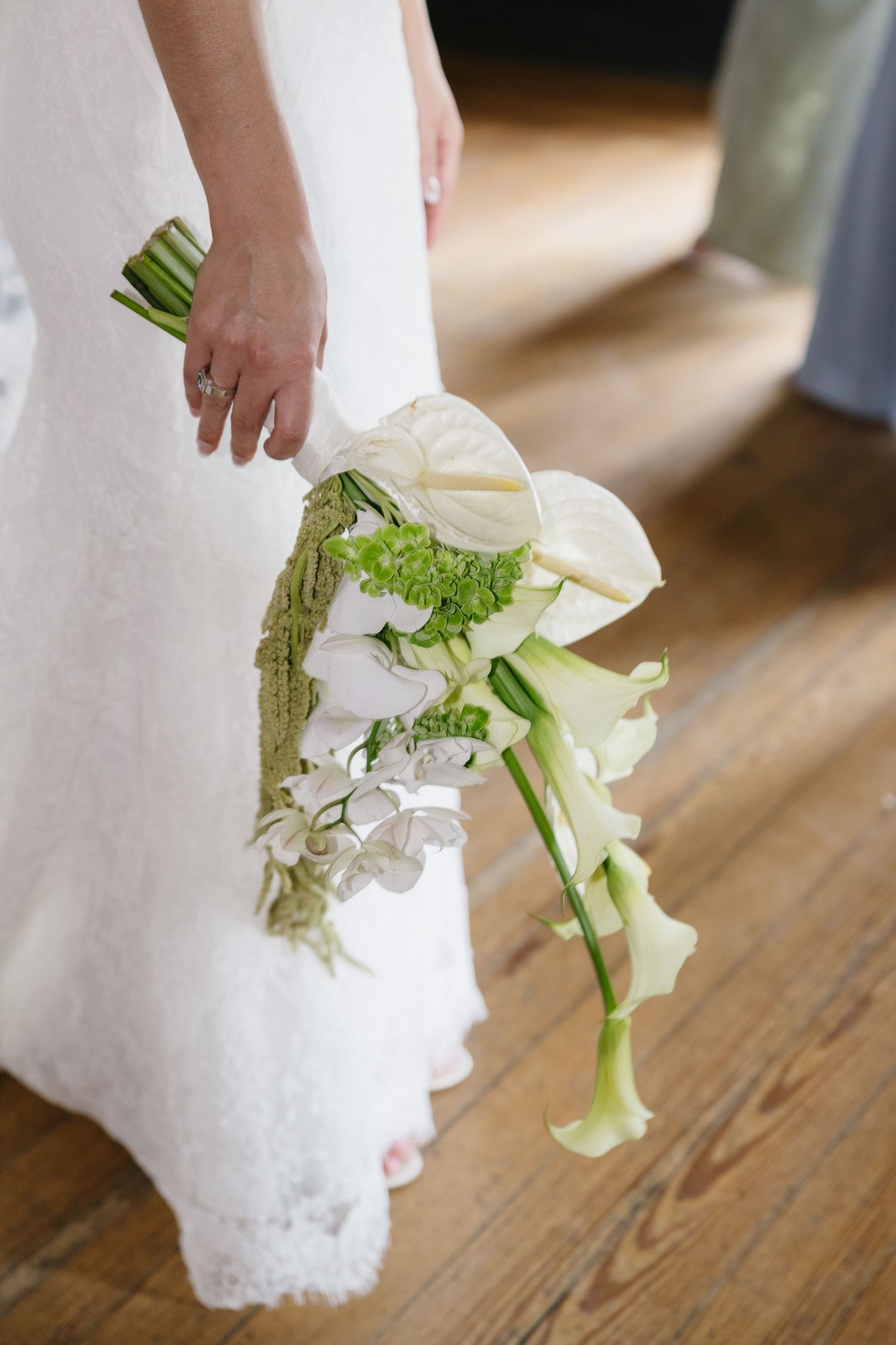 Bride holding cascading white calla lily and anthurium wedding bouquet with green accents against lace gown.