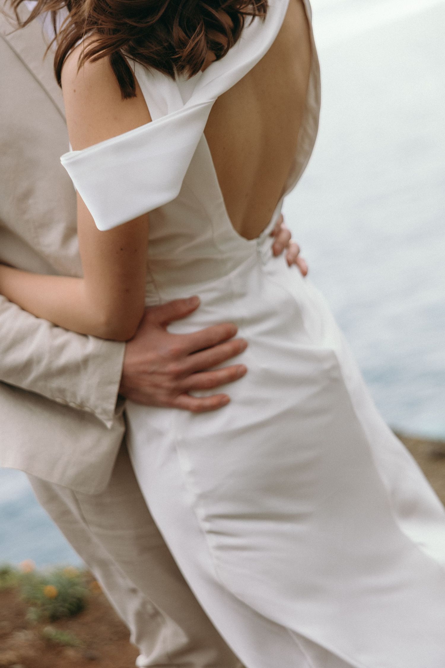 Close-up of groom embracing bride in an elegant open-back white wedding dress against an ocean backdrop.