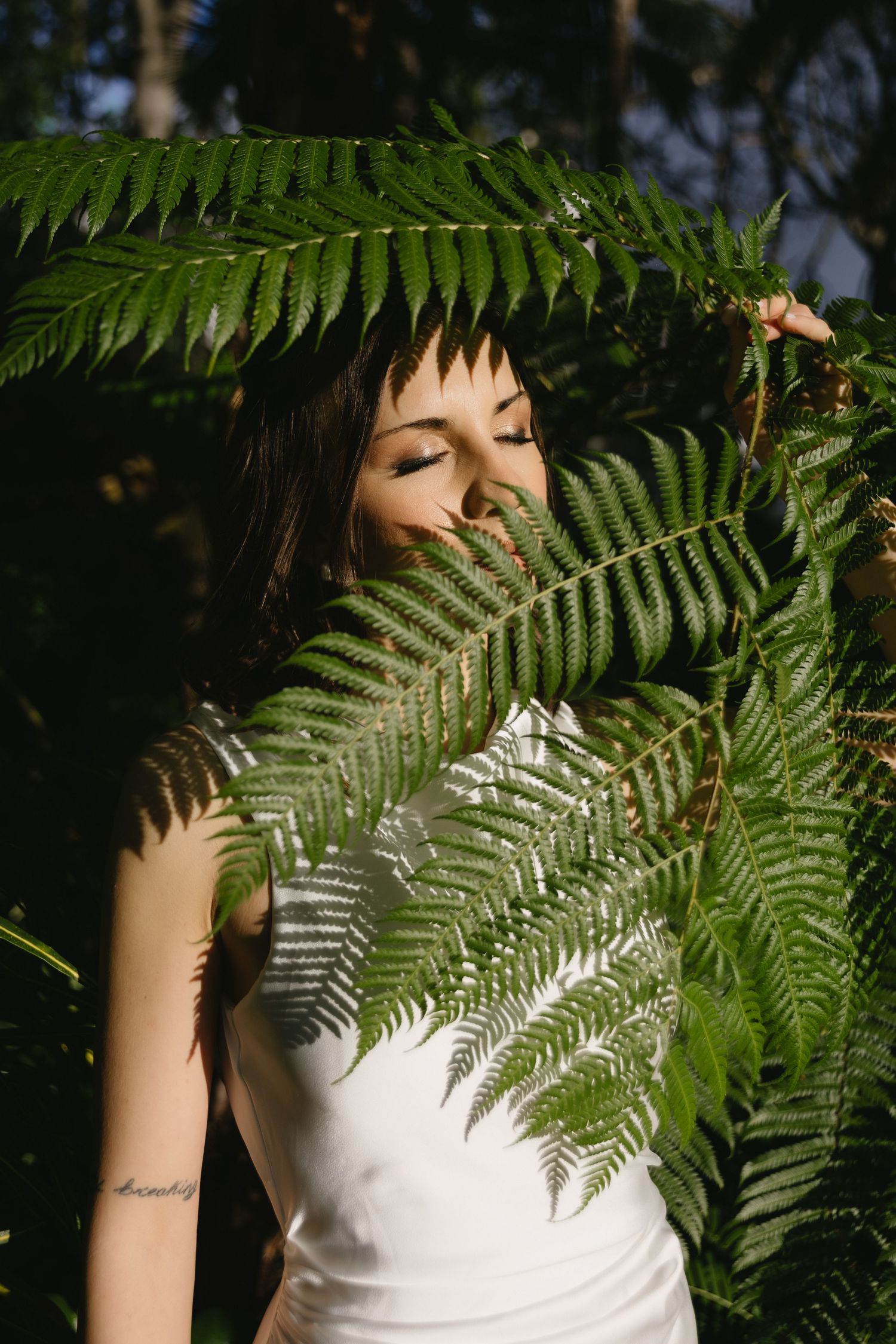 Bride in white dress poses with eyes closed behind large sunlit green fern fronds in forest.