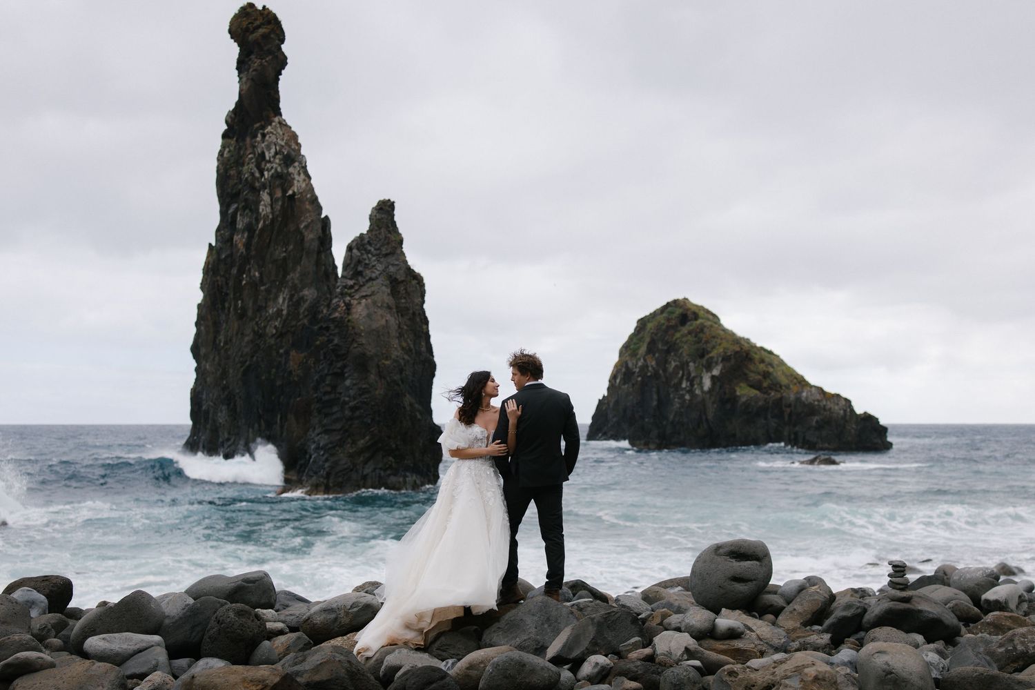 Wedding couple embraces on rocky shoreline with dramatic sea stacks and crashing ocean waves behind them.