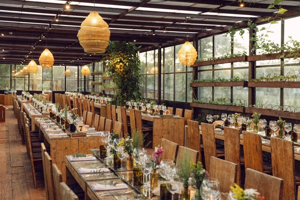 Elegant greenhouse wedding reception with long wooden tables, wicker pendant lights, and lush greenery decor.