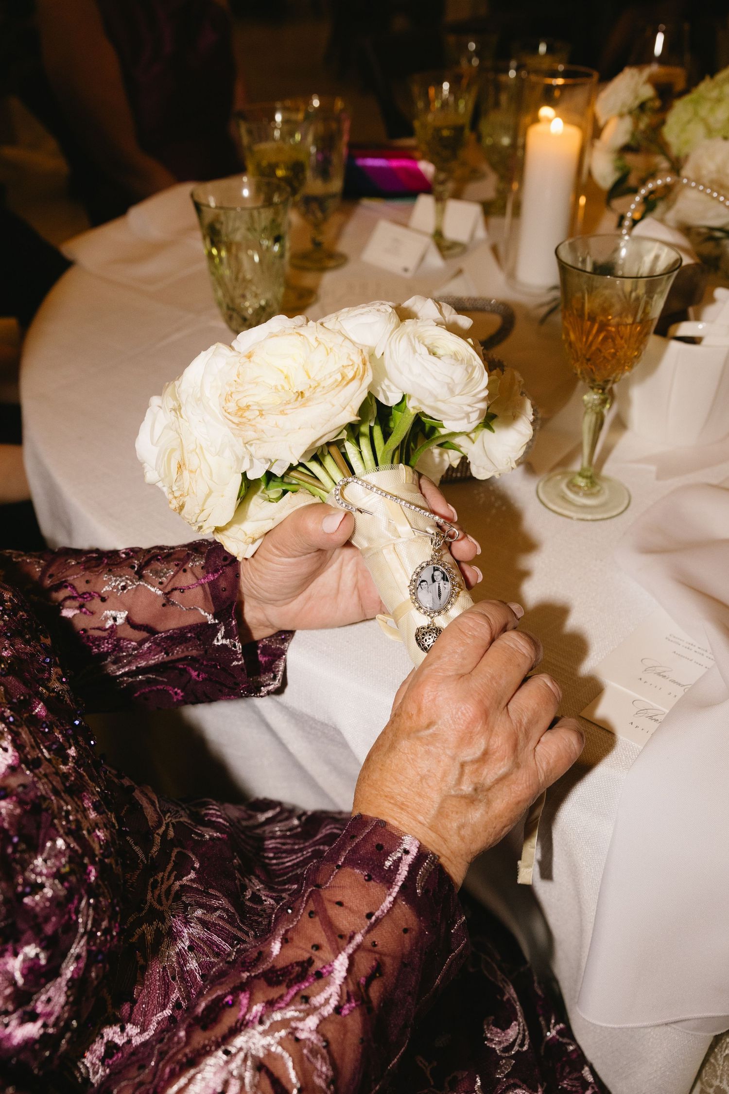 Elderly woman in purple velvet dress holding white rose bouquet with vintage locket charm at wedding reception table.