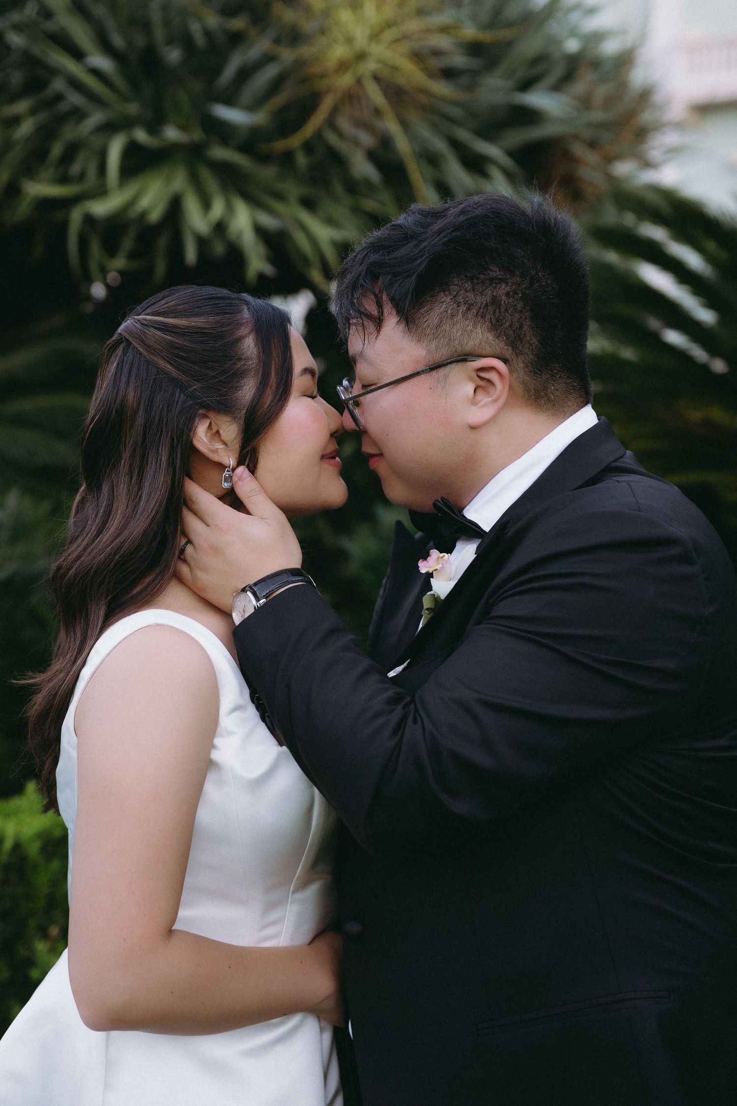 A bride and groom share a romantic almost-kiss moment outdoors, dressed in wedding attire with lush greenery behind them.