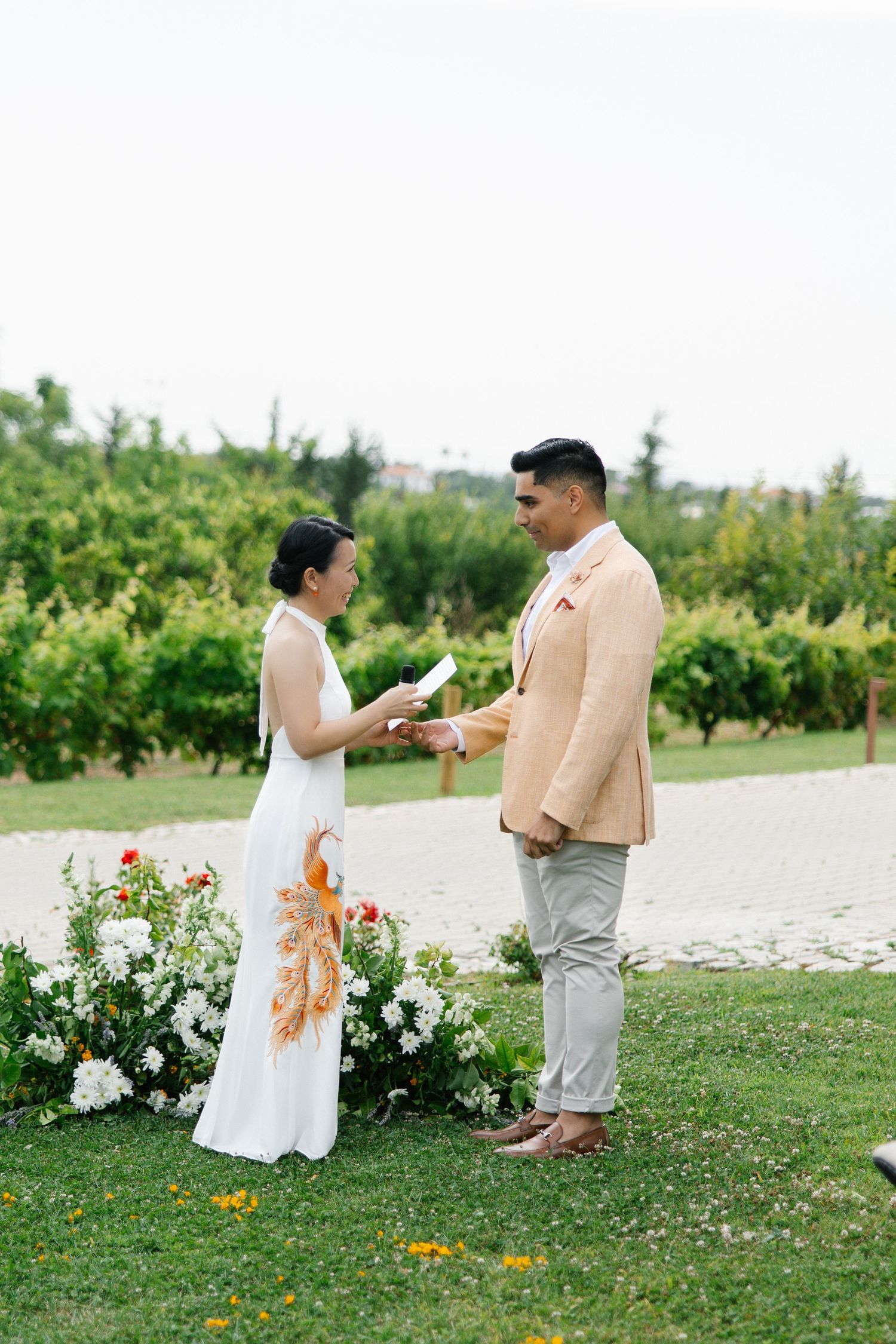 Bride in white phoenix-embroidered dress reads vows to groom in peach blazer during outdoor vineyard wedding ceremony.