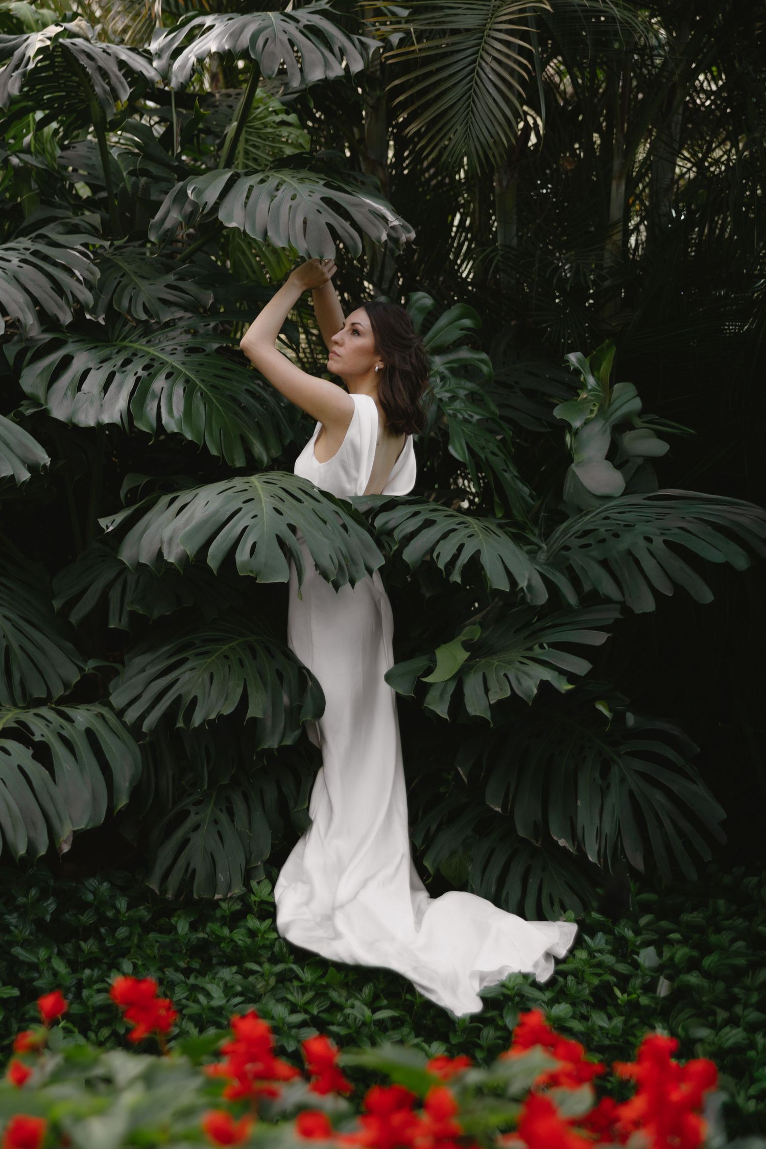 Bride in flowing white gown poses gracefully among large tropical monstera leaves and lush green foliage.