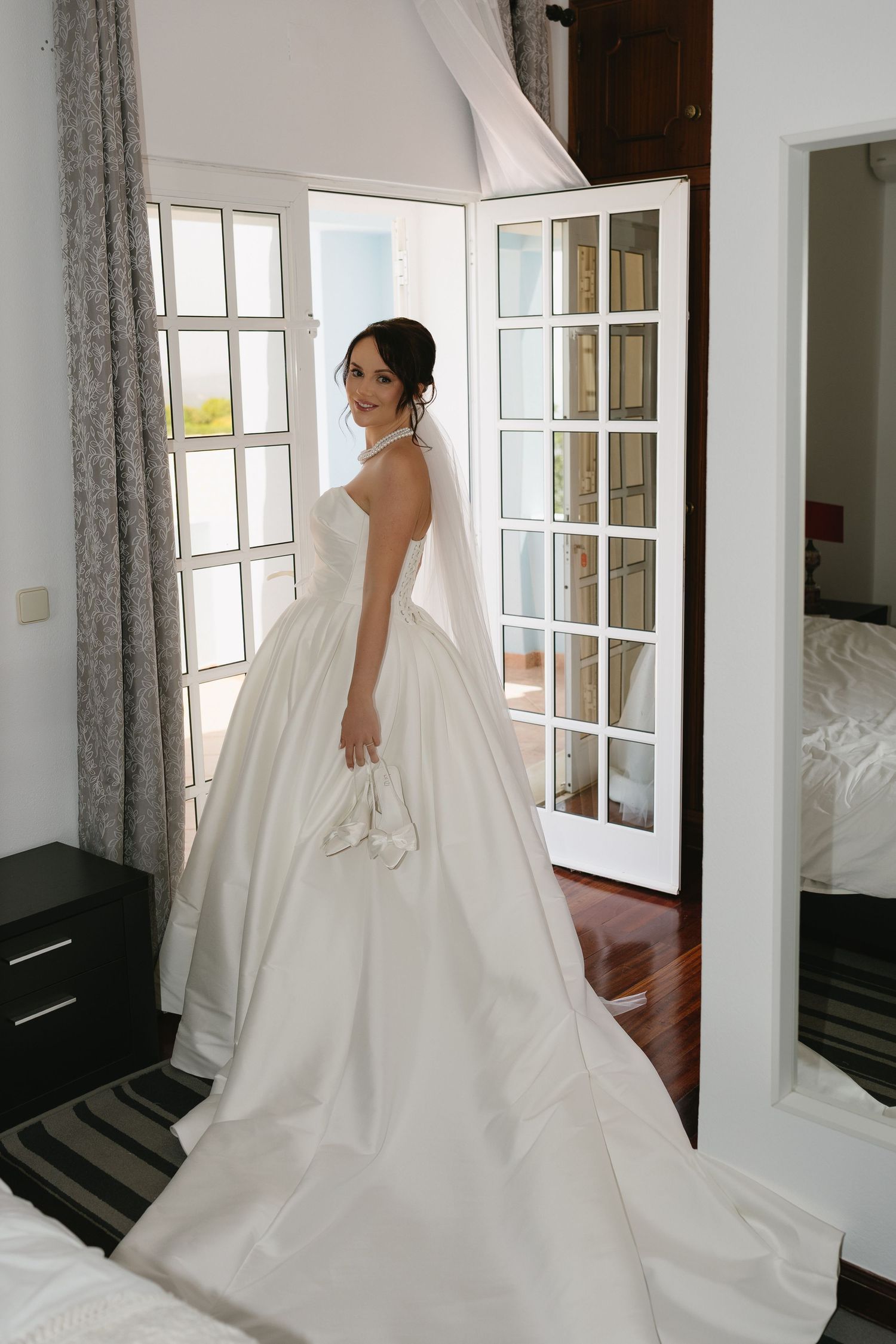 Elegant bride in a white ball gown with veil stands by French doors in a sunlit bridal suite.