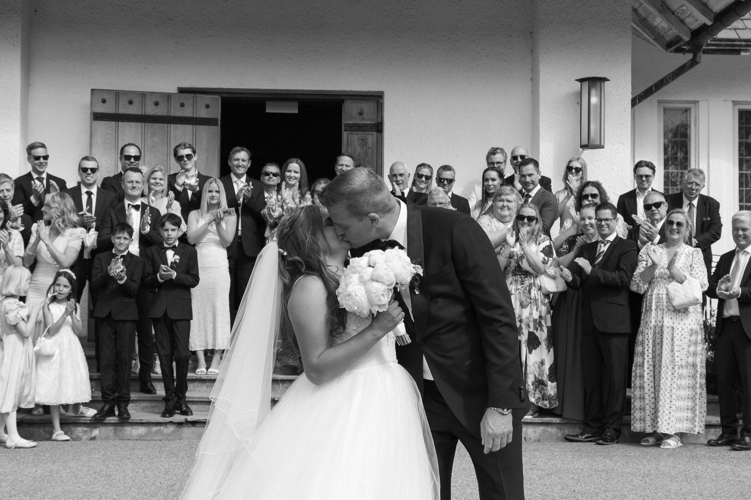 Black and white wedding photo of bride and groom kissing outside church surrounded by applauding guests.
