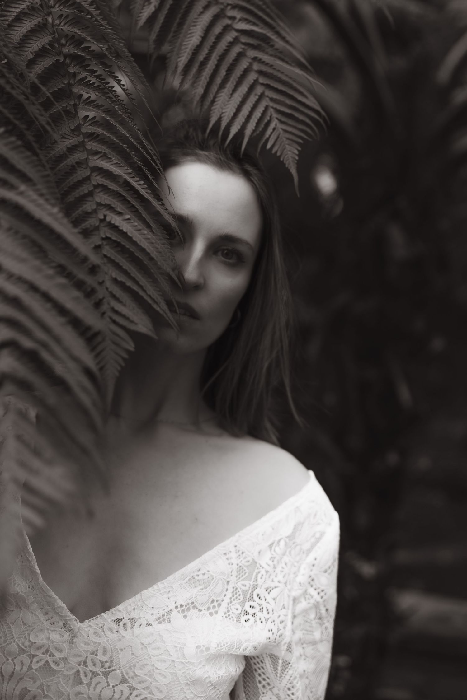 Black and white portrait of a bride in off-shoulder lace gown surrounded by lush tropical fern leaves.