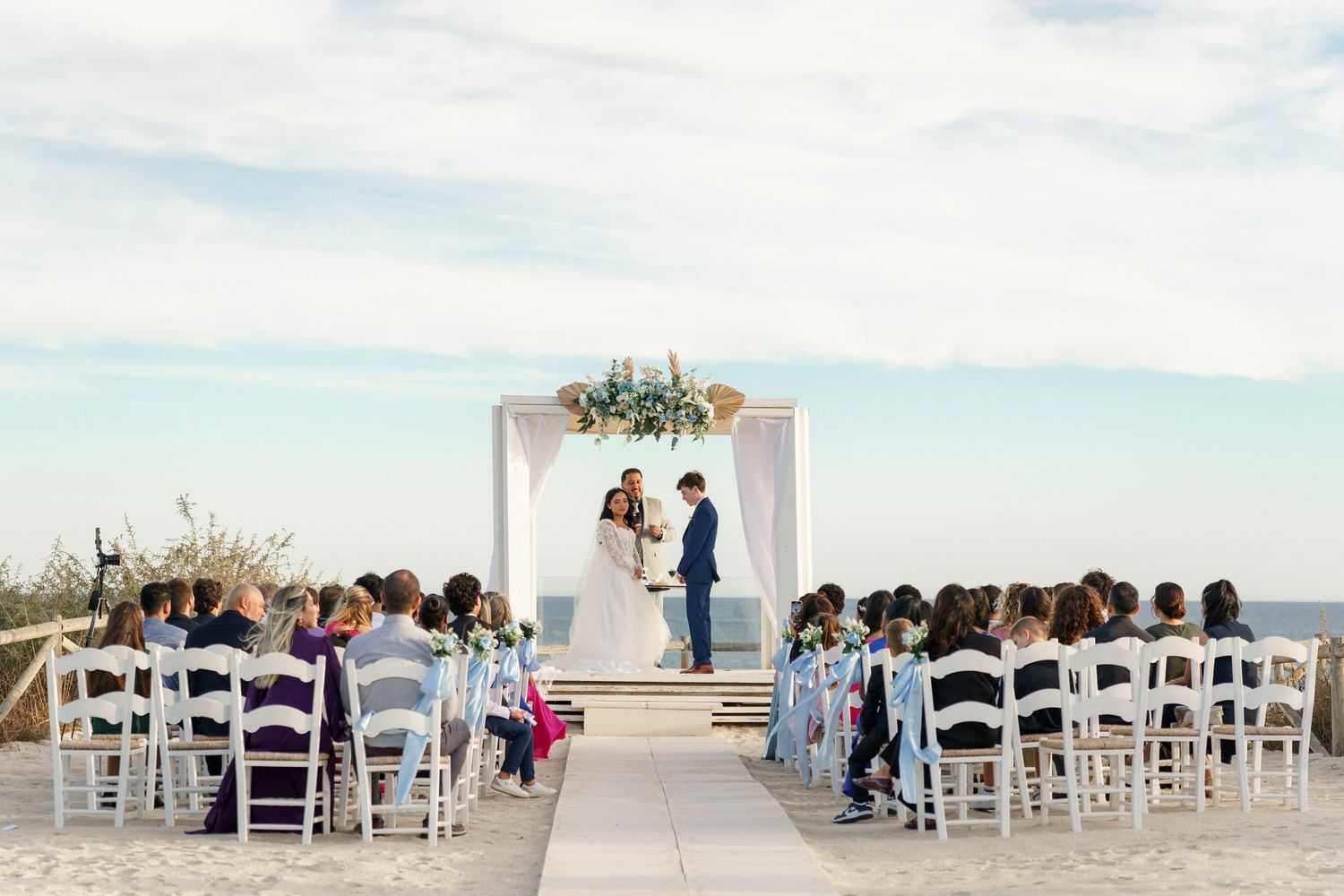 Couple exchanging vows at a white arch wedding ceremony on the beach with seated guests.