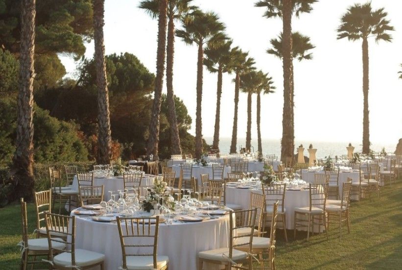 Elegant outdoor wedding reception with round white-draped tables, gold chiavari chairs, and tall palm trees at sunset.