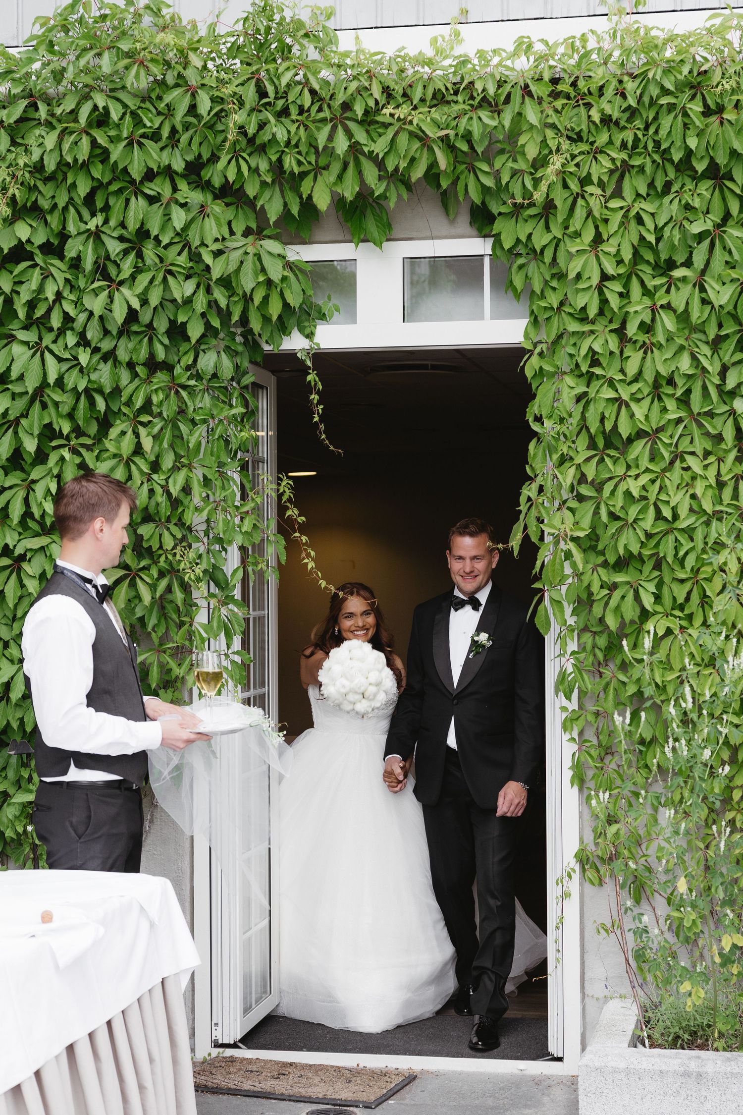 Bride and groom exit through ivy-covered doorway as a waiter holds champagne at an elegant outdoor wedding venue.