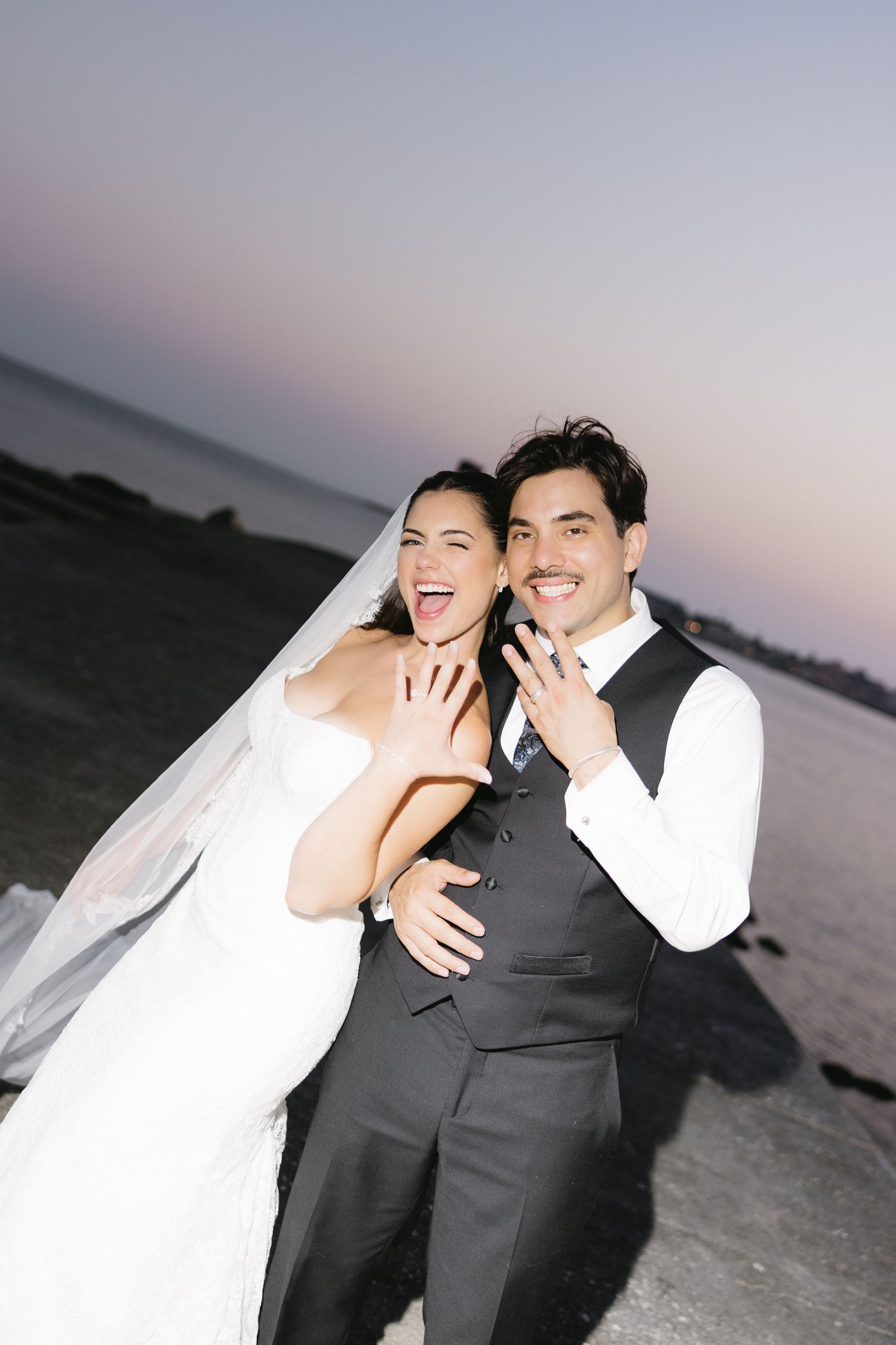 Happy newlyweds showing wedding rings at sunset by the sea, bride in white veil and groom in gray suit smiling joyfully.