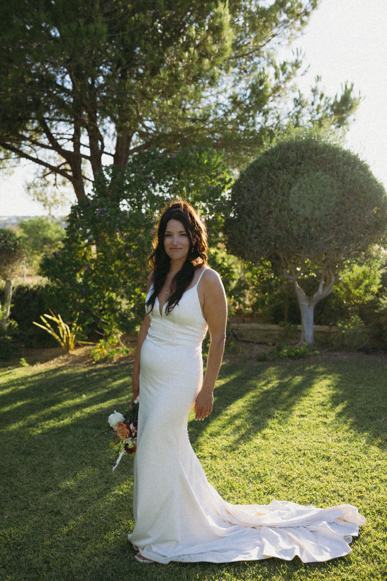 Bride posing on green grass with lush green trees in the background during wedding photoshoot