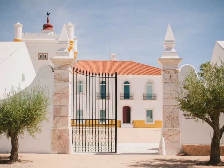 White Portuguese villa with ornate iron gates, terracotta roof, and olive trees under a clear blue sky.