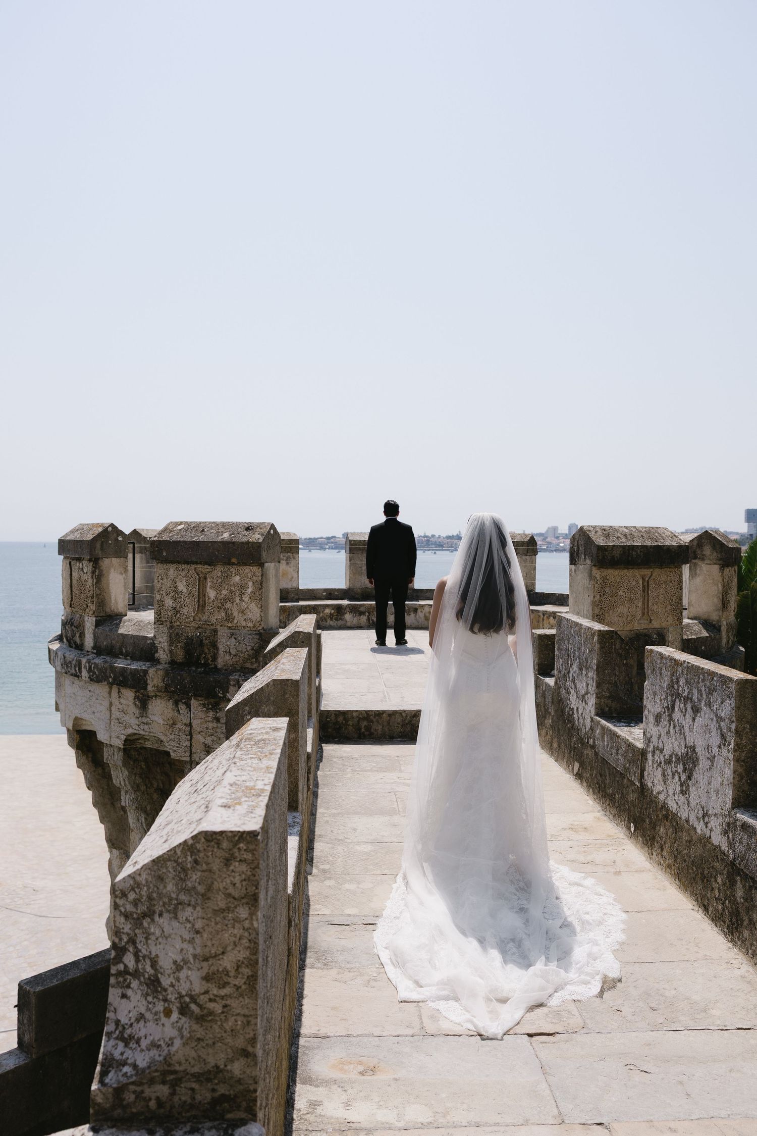 Bride in white gown and veil walks toward groom on stone castle wall overlooking the ocean.
