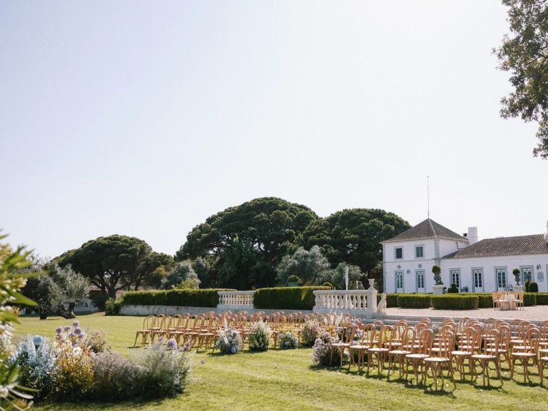 Outdoor wedding ceremony with wooden chairs on a green lawn beside a classic white manor house and gardens.