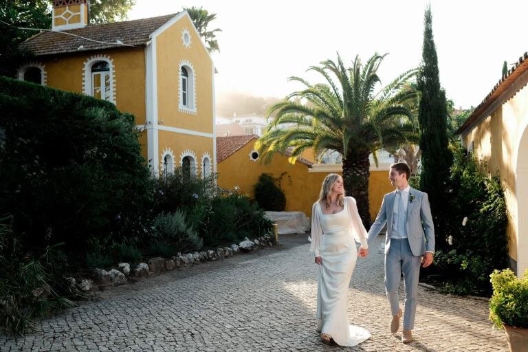 Bride and groom strolling along a cobblestone path beside a vibrant yellow villa with palm trees.