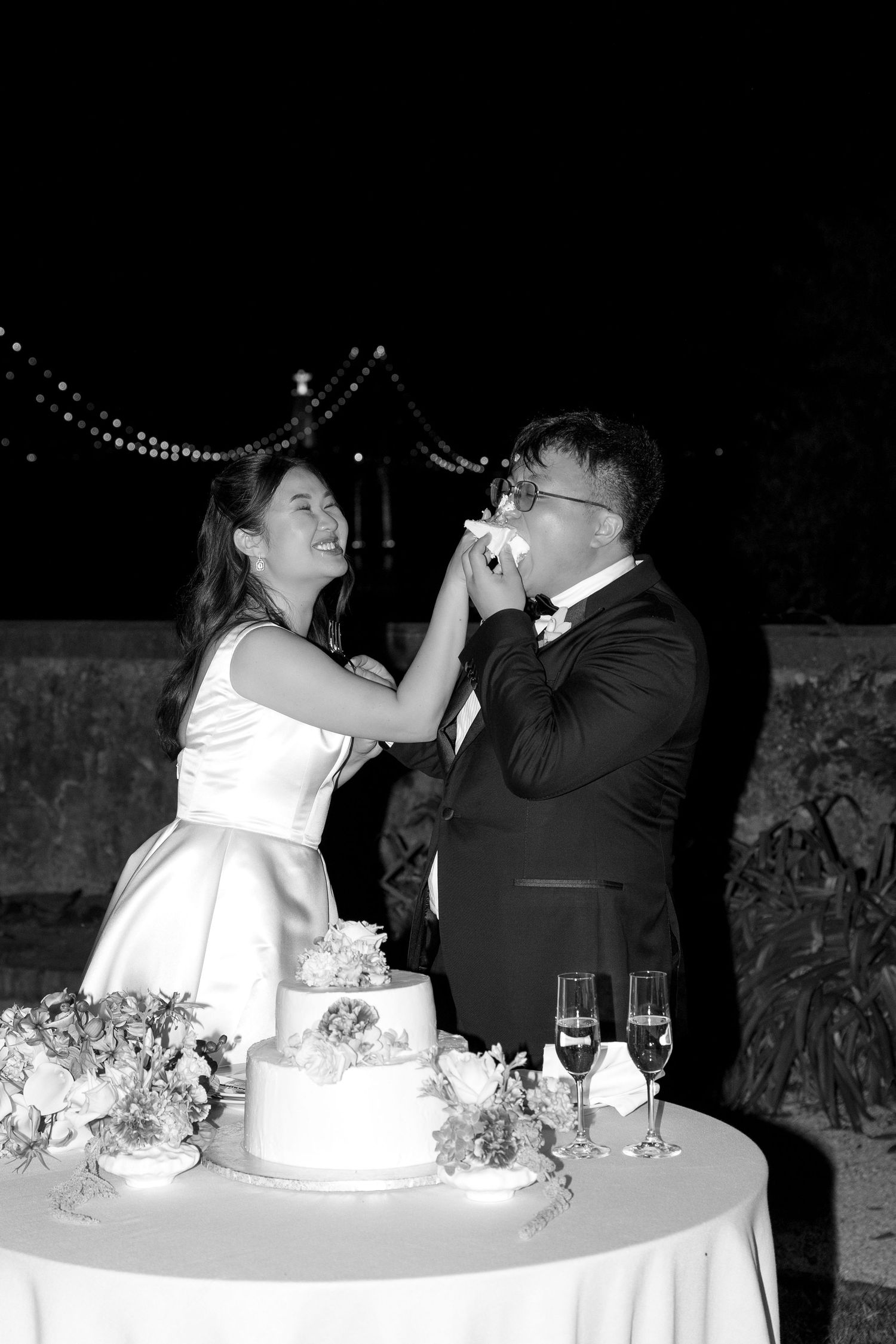 Black and white photo of bride feeding groom wedding cake at outdoor evening reception with champagne glasses nearby.