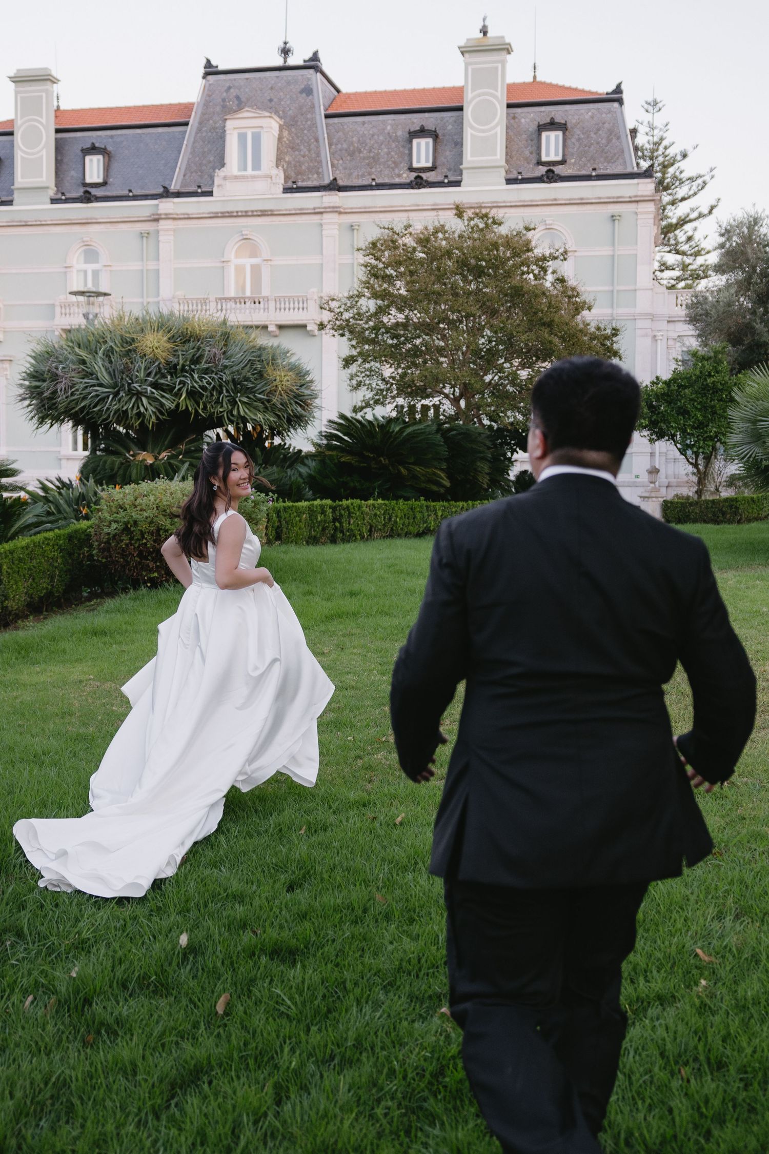 Bride in white gown and groom in black suit running toward each other on green lawn before elegant mansion.