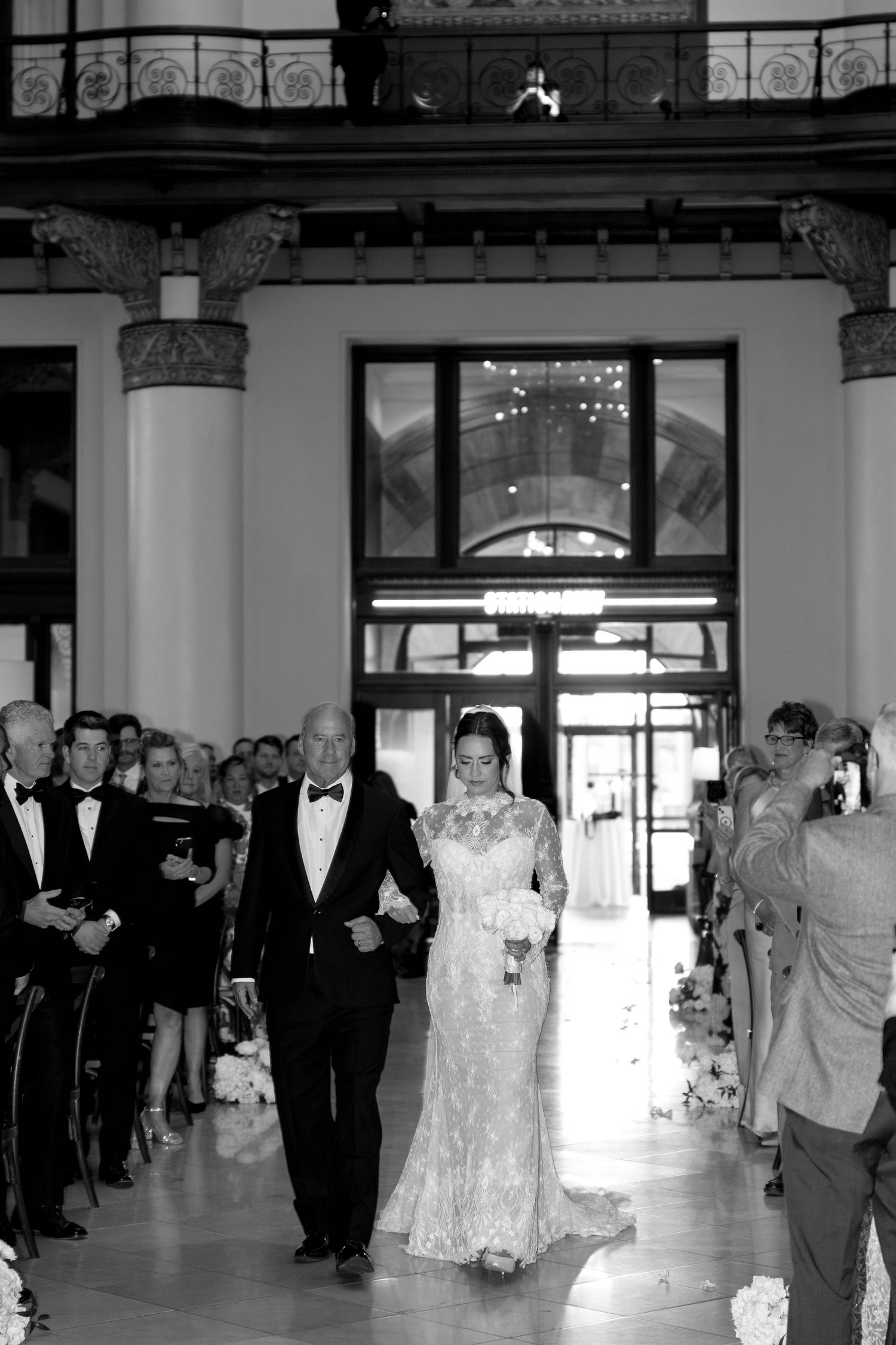 Black and white photo of a bride in lace gown walking down the aisle with her father at an elegant wedding ceremony.