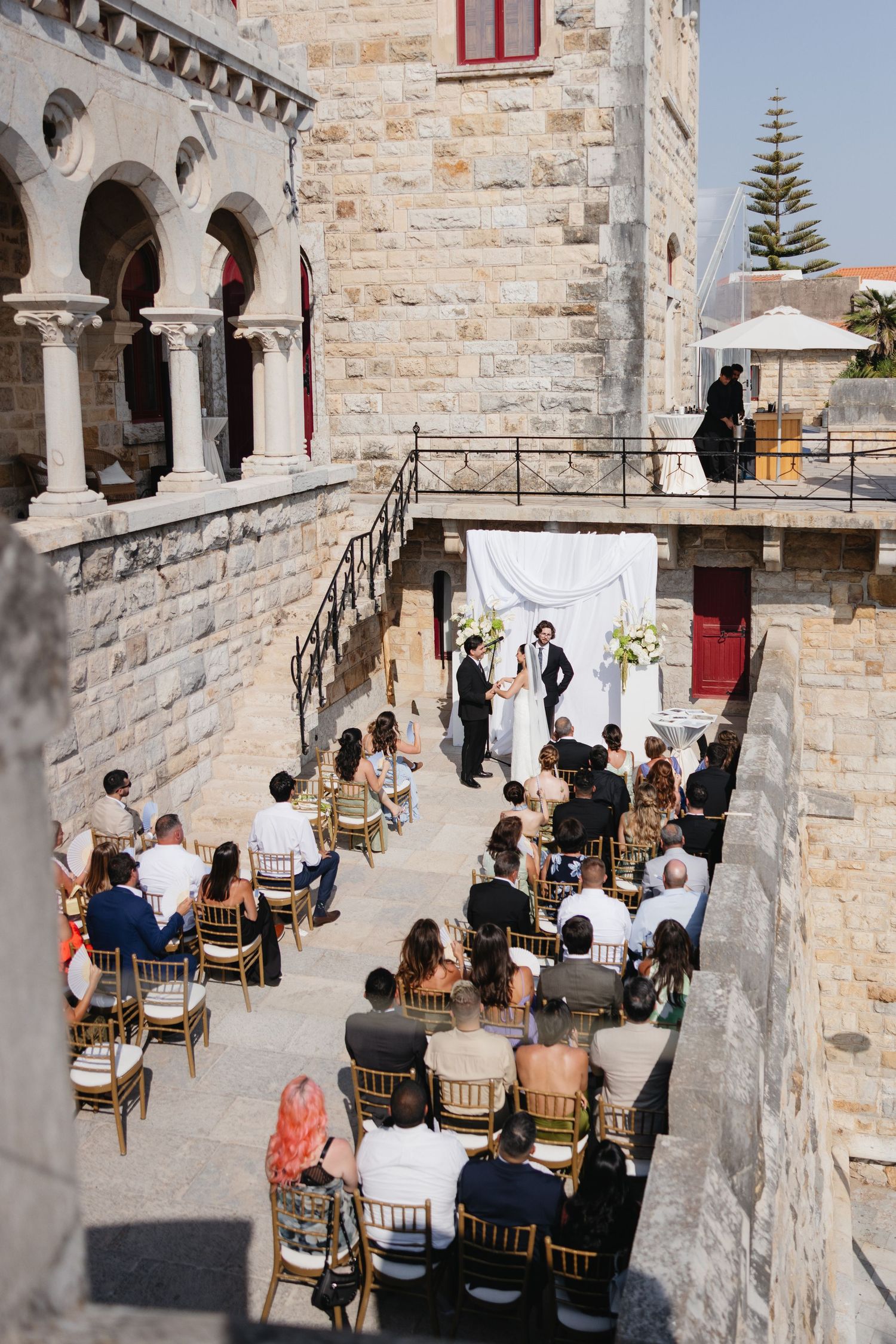 Outdoor wedding ceremony at a historic stone castle venue with guests seated in gold chairs facing the altar.