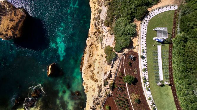 Breathtaking aerial view of cliffside wedding venue with turquoise ocean waters and white ceremony setup on green terrace.