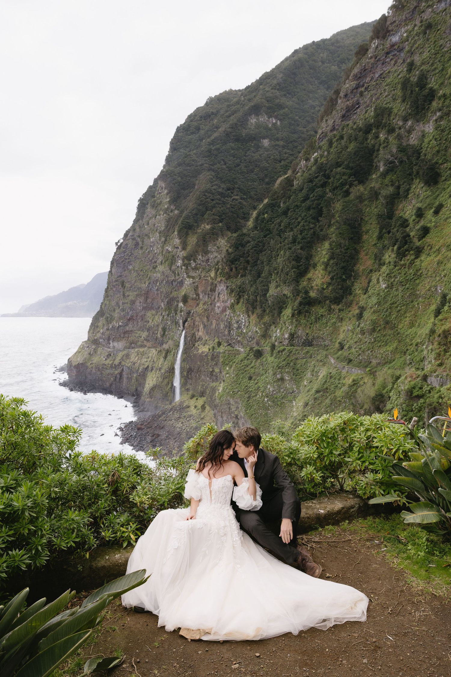 Bride and groom sit intimately on a cliffside ledge with a waterfall and ocean visible in the background.