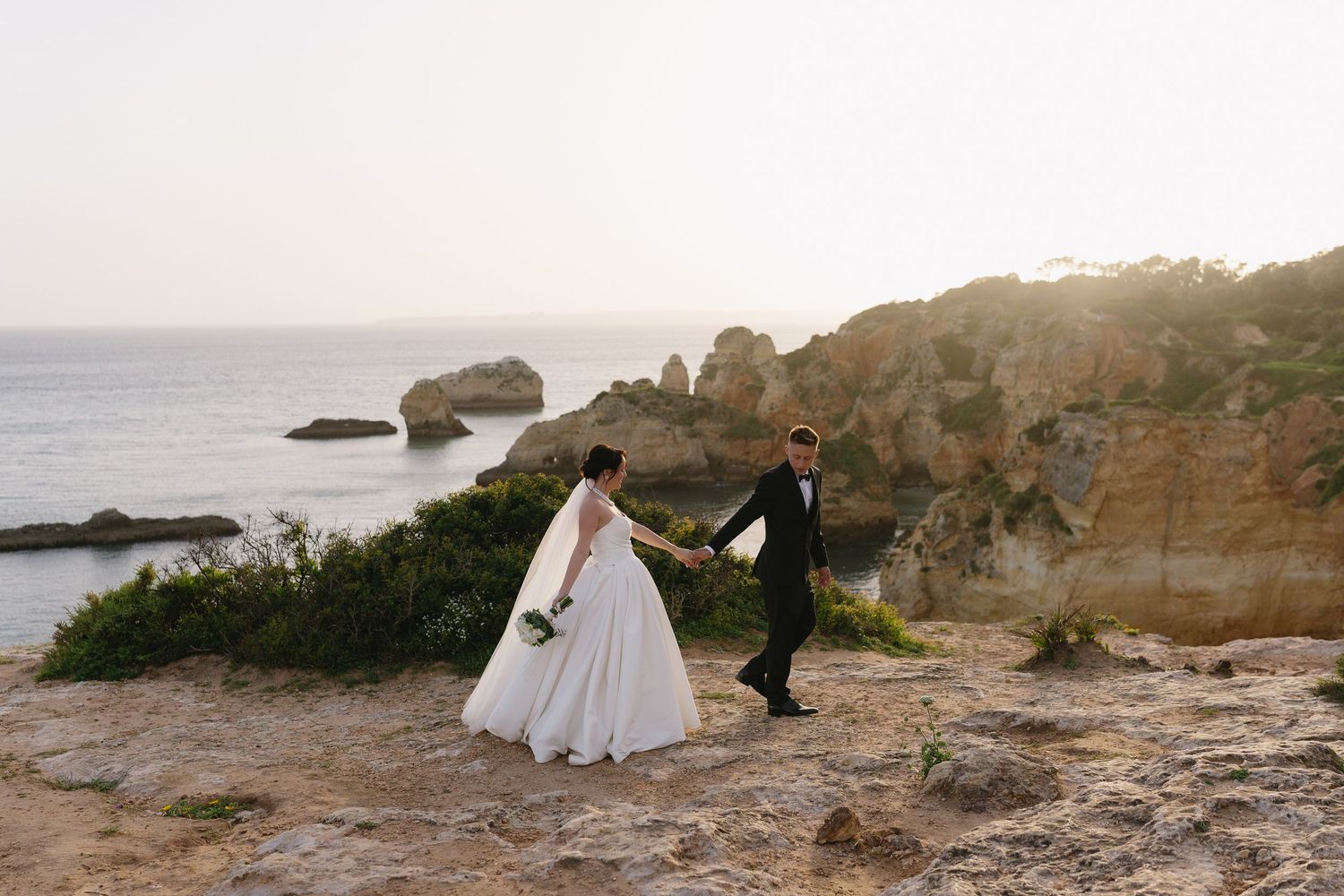 Bride and groom holding hands strolling along scenic rocky coastal cliffs during a golden sunset.