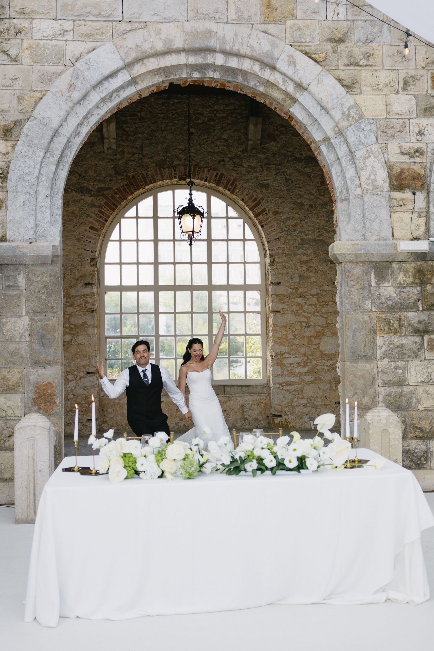 Bride and groom celebrate at elegant sweetheart table with white florals beneath stone arch at wedding venue.