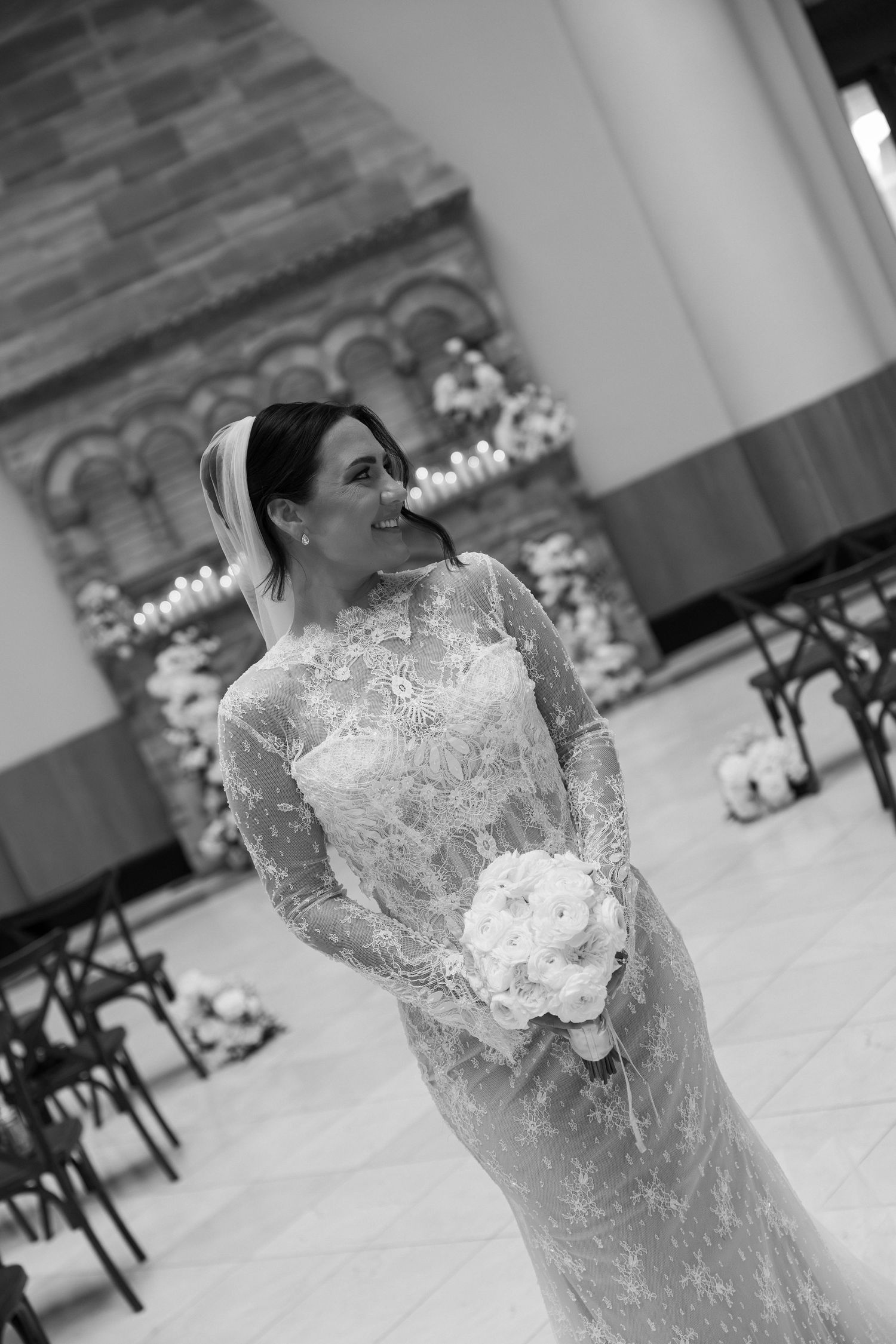 Bride in lace long-sleeve wedding gown holding white bouquet, smiling in elegant venue aisle, black and white photo.