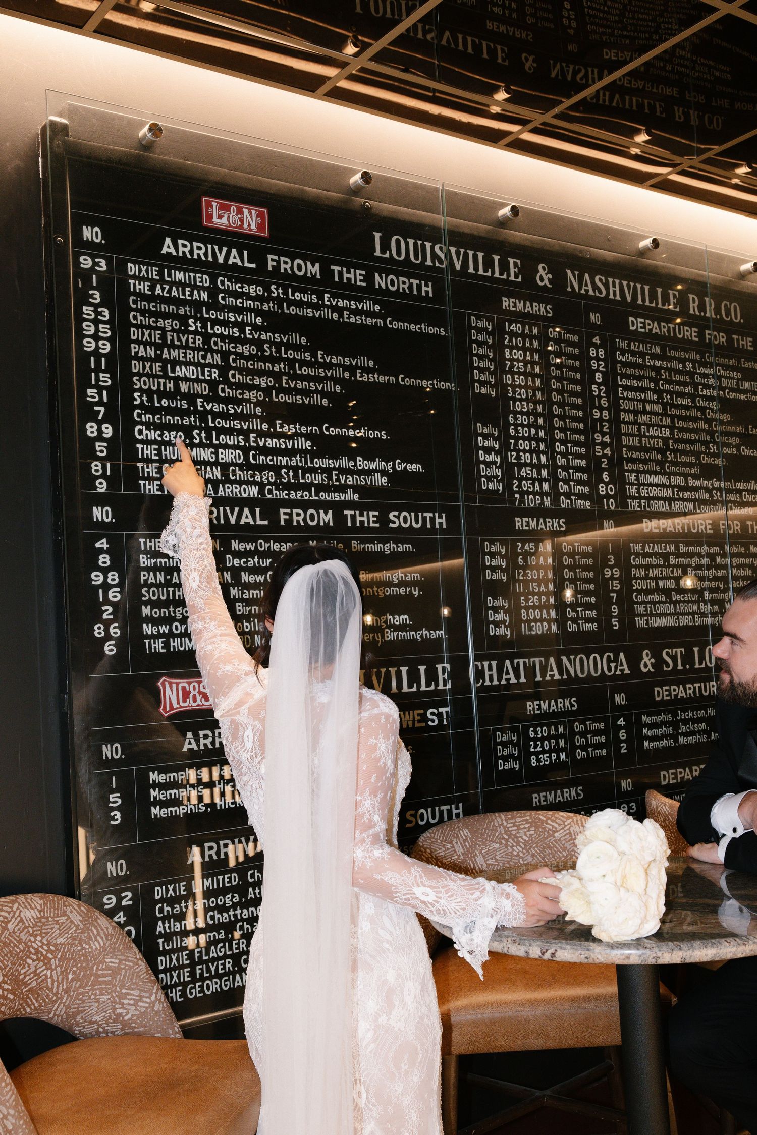 Bride in lace gown and veil points at Louisville & Nashville Railroad departure board at wedding venue.