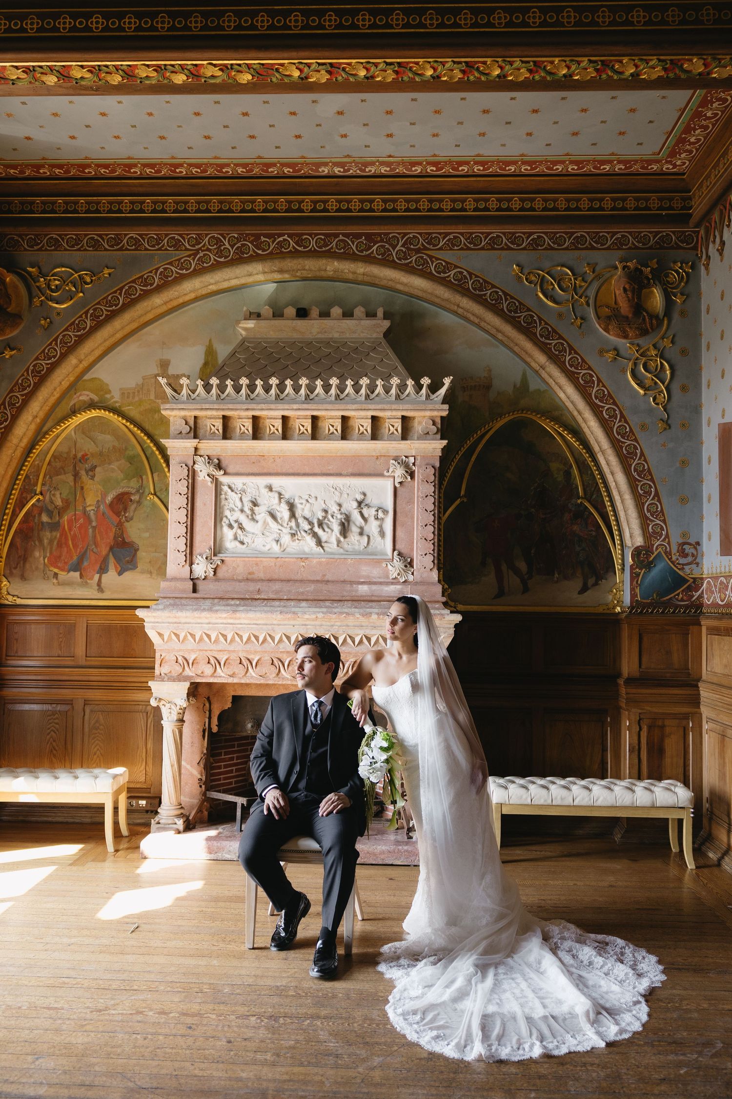 Bride in lace gown and groom in gray suit pose in ornate room with gilded arches and decorative fireplace.