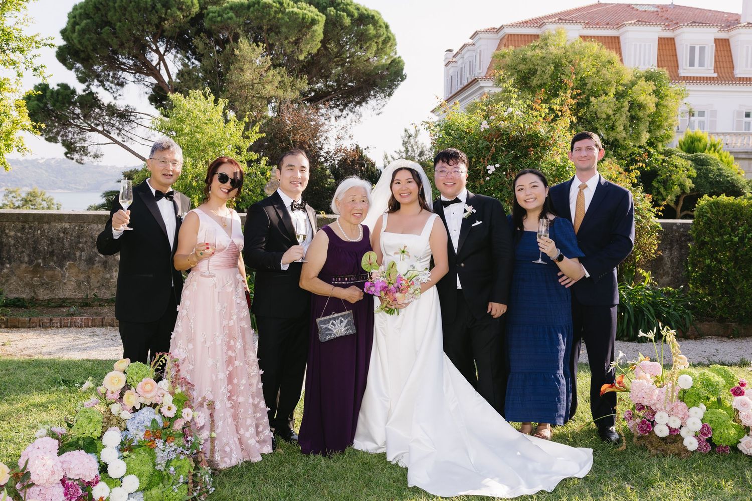 Wedding party poses outdoors in formal attire, bride in white gown holding bouquet, surrounded by family at elegant garden venue.