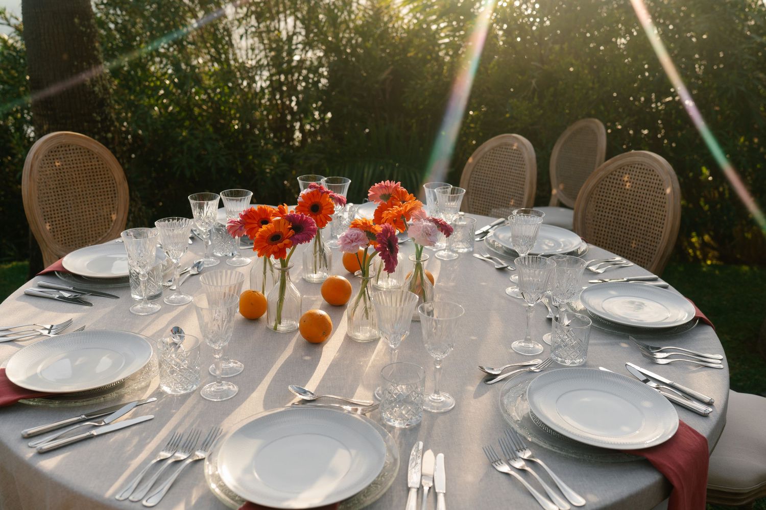 Elegant outdoor round dining table set with crystal glasses, white plates, and vibrant orange and red floral centerpieces.
