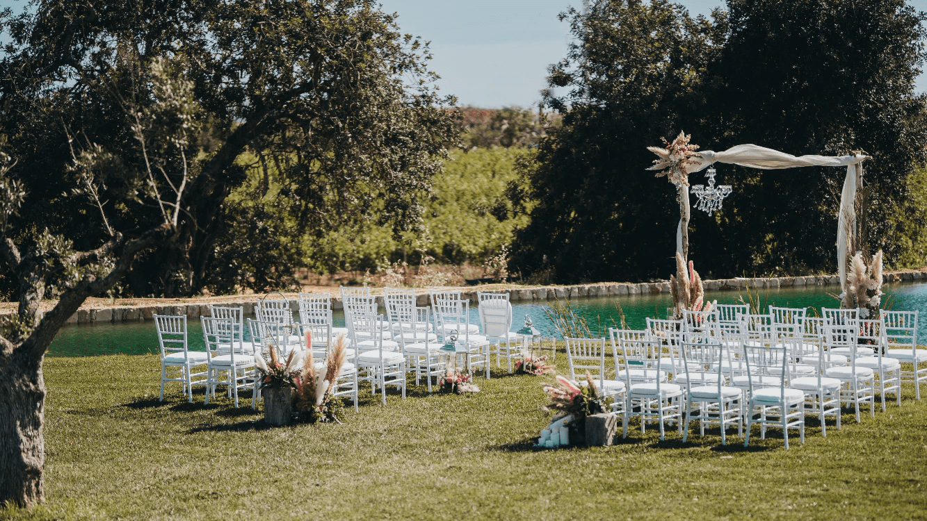 Outdoor wedding ceremony setup on green lawn with white chairs, floral arch, and natural landscape backdrop.