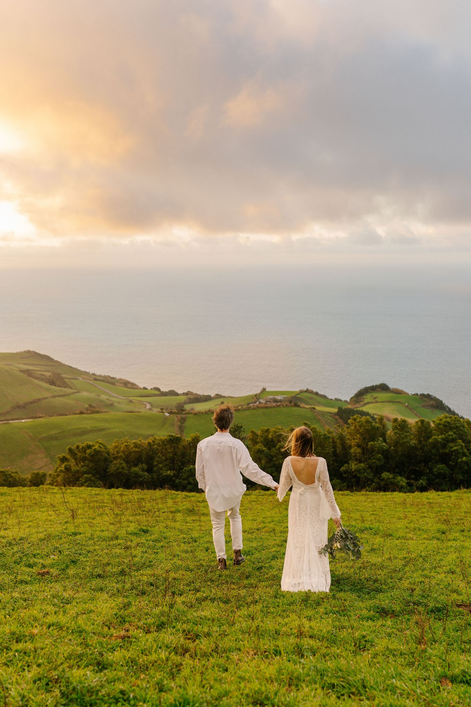 Bride and groom in white attire walk hand-in-hand across green hillside overlooking ocean at dramatic sunset.