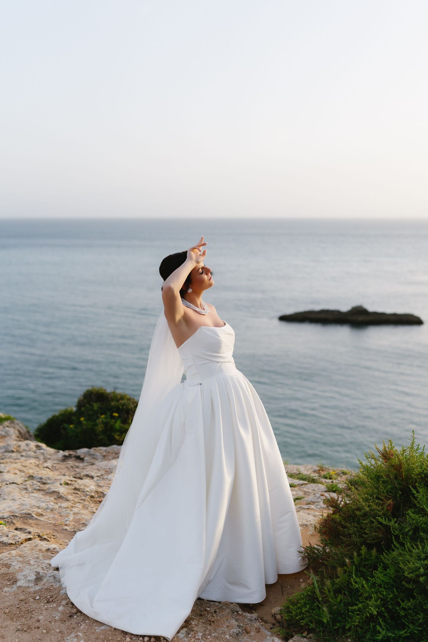 Bride in strapless ballgown with long veil posing on coastal cliff with serene blue ocean backdrop.