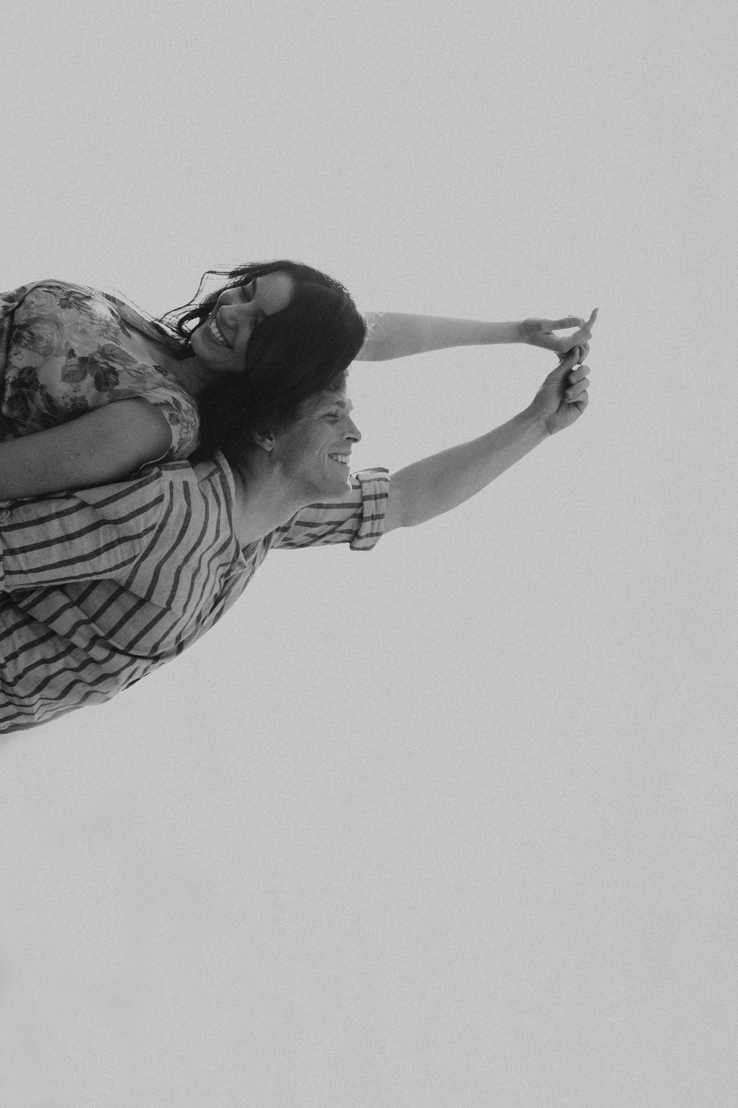 Black and white candid shot of a joyful couple, woman riding piggyback, both laughing against a light sky.