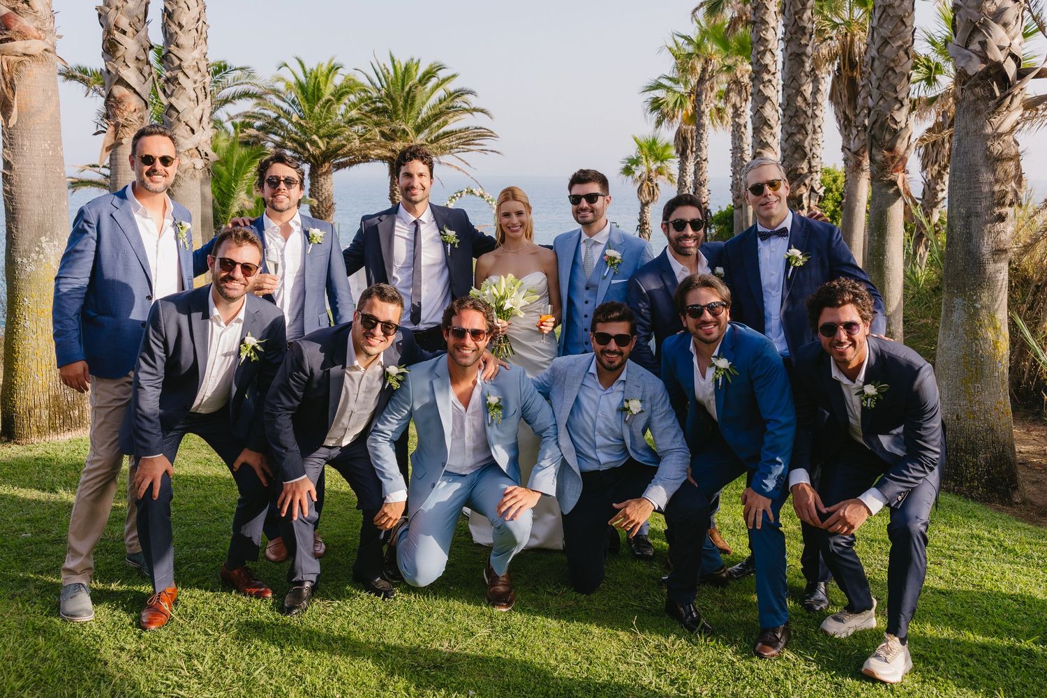 Groom poses with groomsmen in blue suits on a green lawn with palm trees at an outdoor wedding.