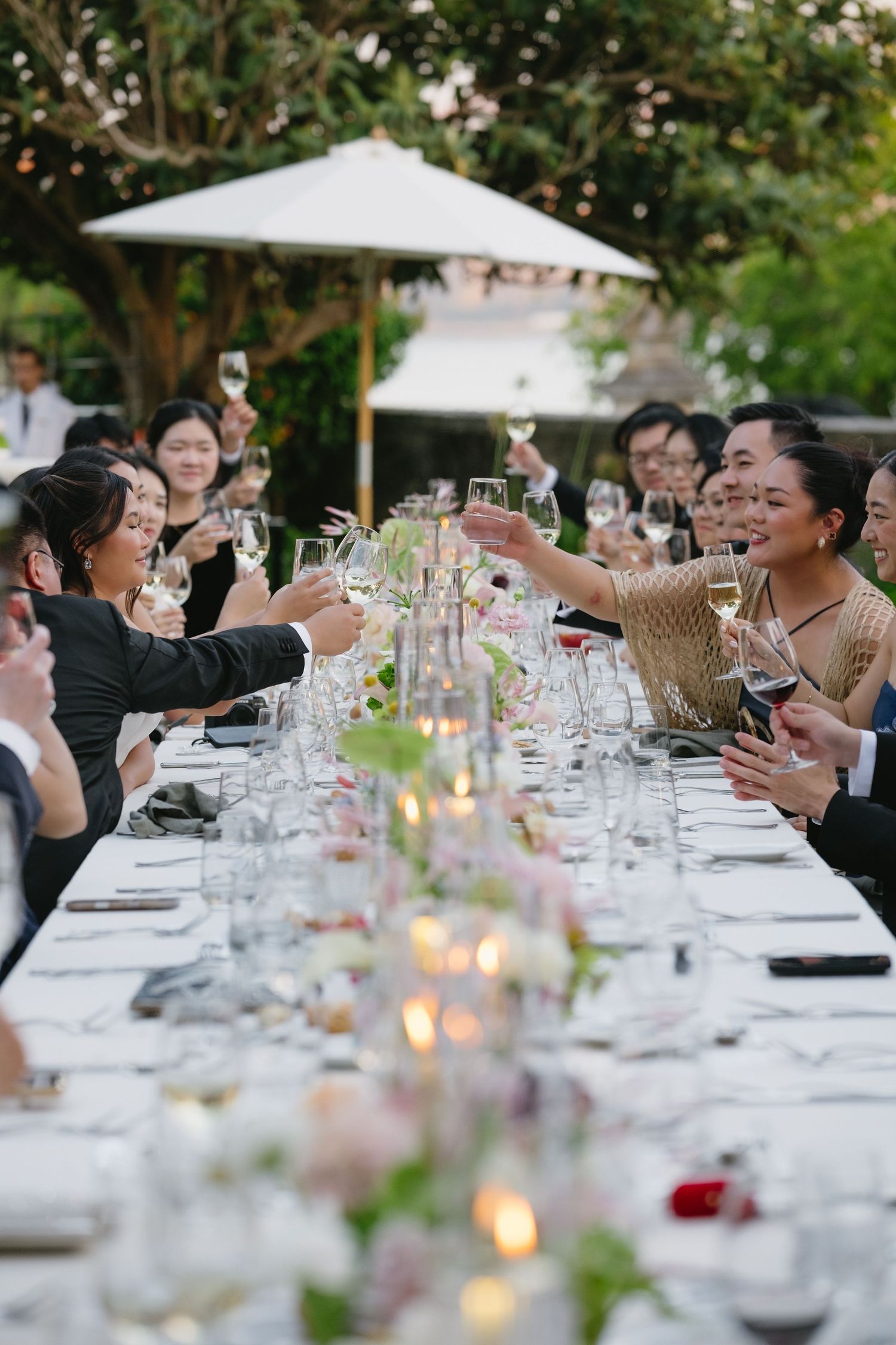 Guests raising wine glasses in a toast at an elegant outdoor wedding reception dinner with candles and floral centerpieces.